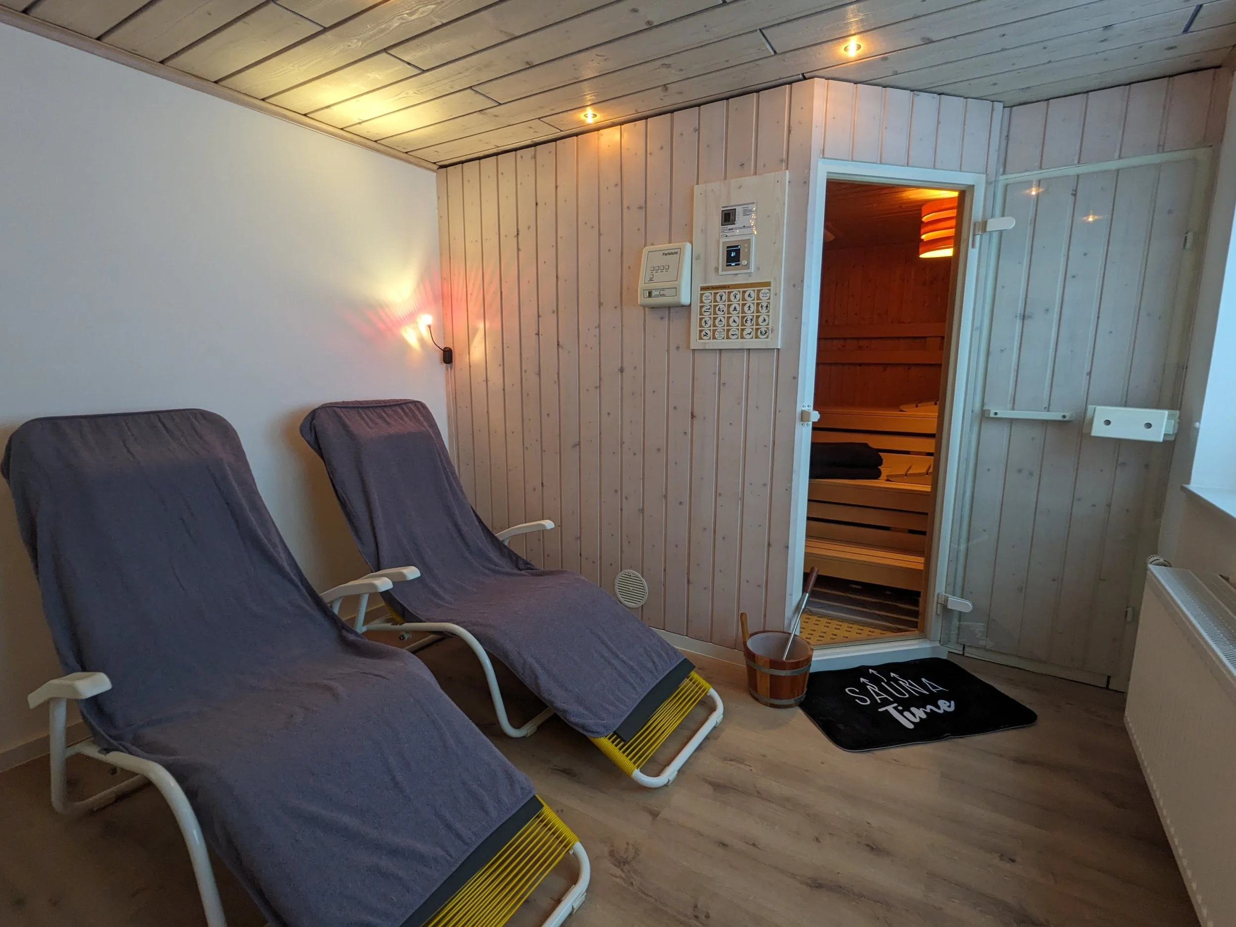 Drei Liegestühle stehen vor einer weißen Wand. In einem Raum mit Holzdecken und Holzpaneelen ist eine Saunatür sichtbar, die in einen Saunaraum führt. Neben der Tür hängt ein Wandtelefon und eine Steuerungseinheit für die Sauna.