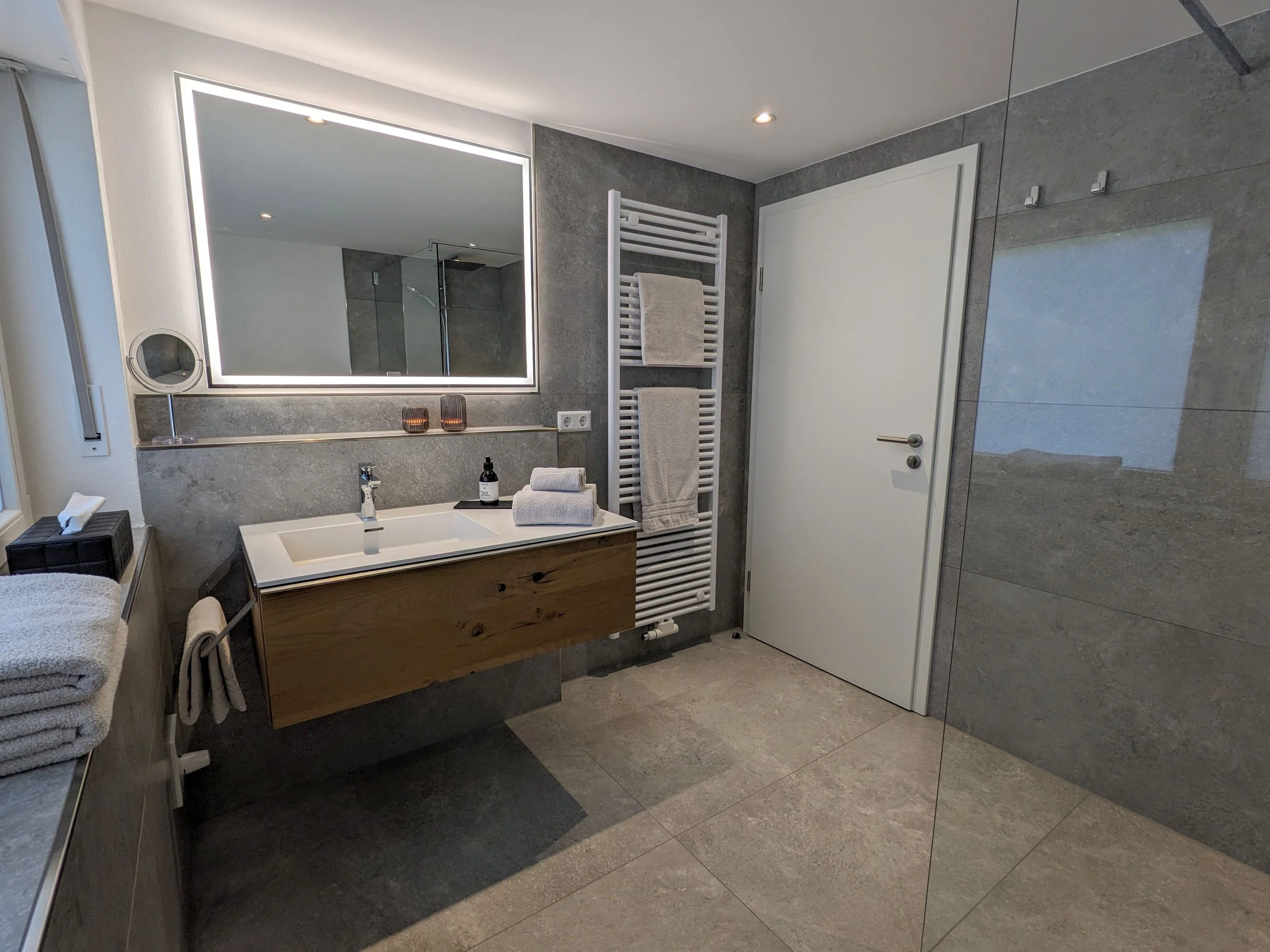 moderner Badezimmer mit großem beleuchteten Spiegel, Waschbecken, Handtuchheizung, weißen Türen und grauen Wand- und Bodenfliesen