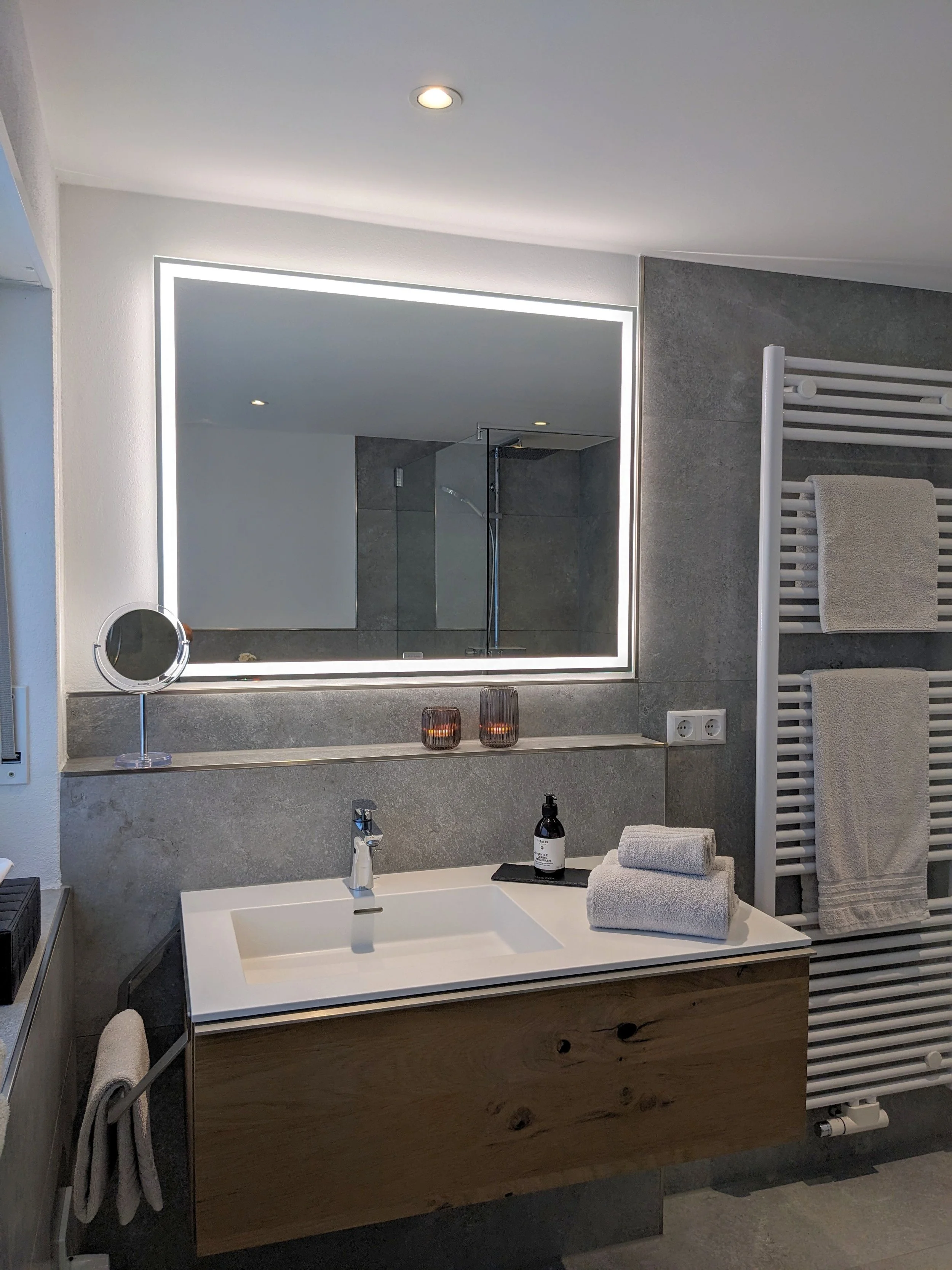 Ein modernes Badezimmer mit einem weißen Waschtisch, grauem Fliesenhintergrund und Handtüchern auf einem Haken, einer beleuchteten Spiegel, Kerzen und einem Waschmittel auf dem Waschtisch.