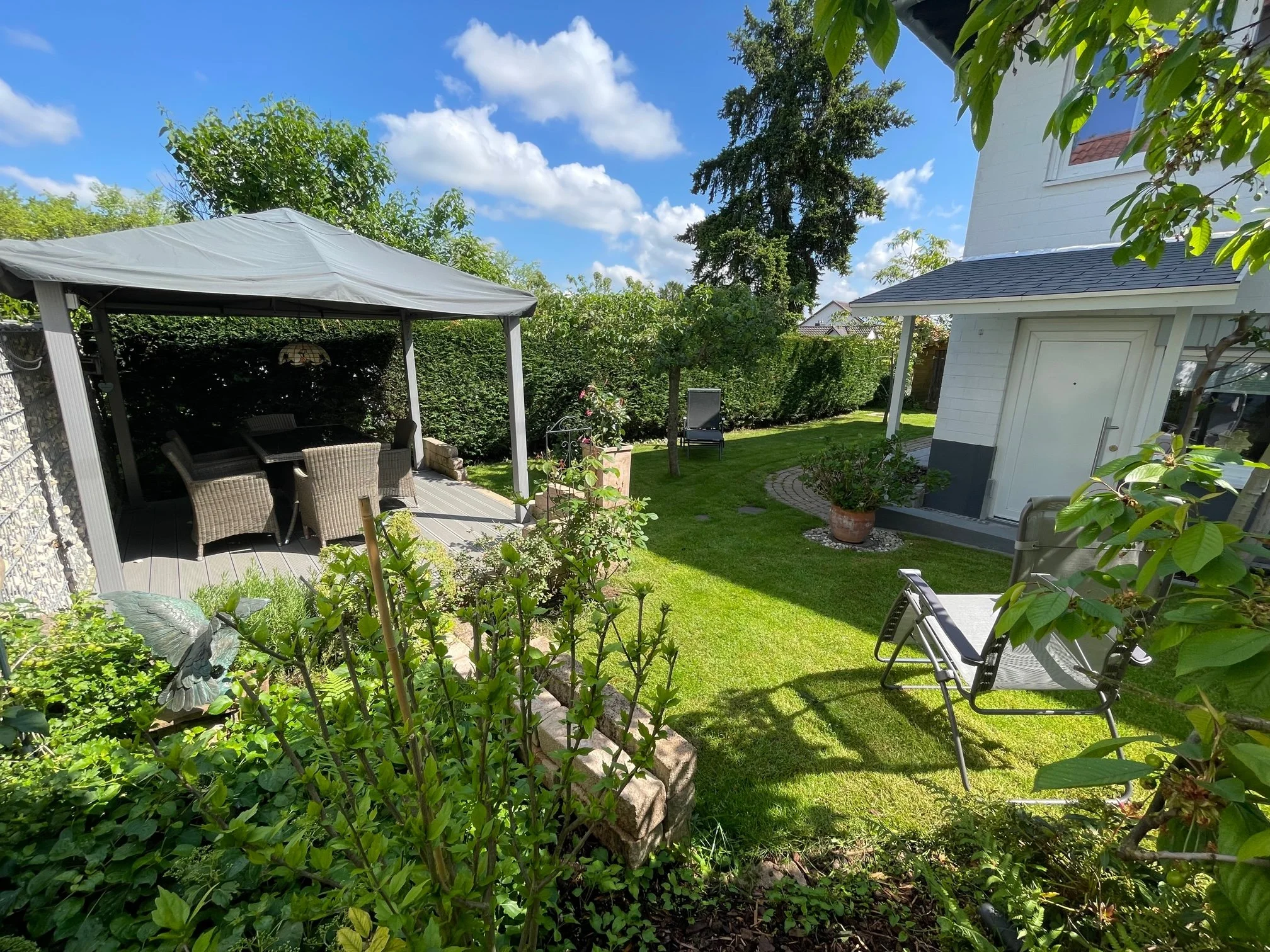 Garten mit Sitzecke unter Pavillon, Rasenfläche, Gartenmöbel und Bäume bei sonnigem Wetter.
