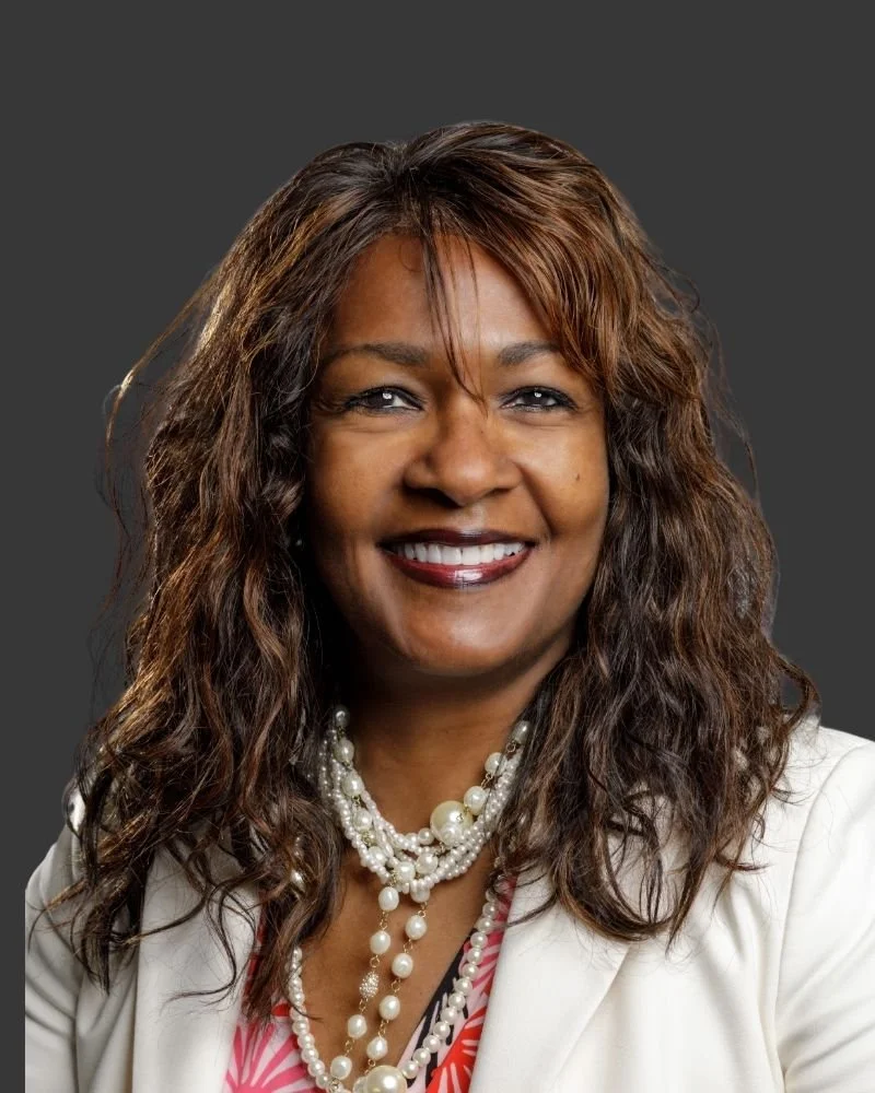 A professional woman with curly hair wearing a white blazer and multiple pearl necklaces, smiling against a gray background.