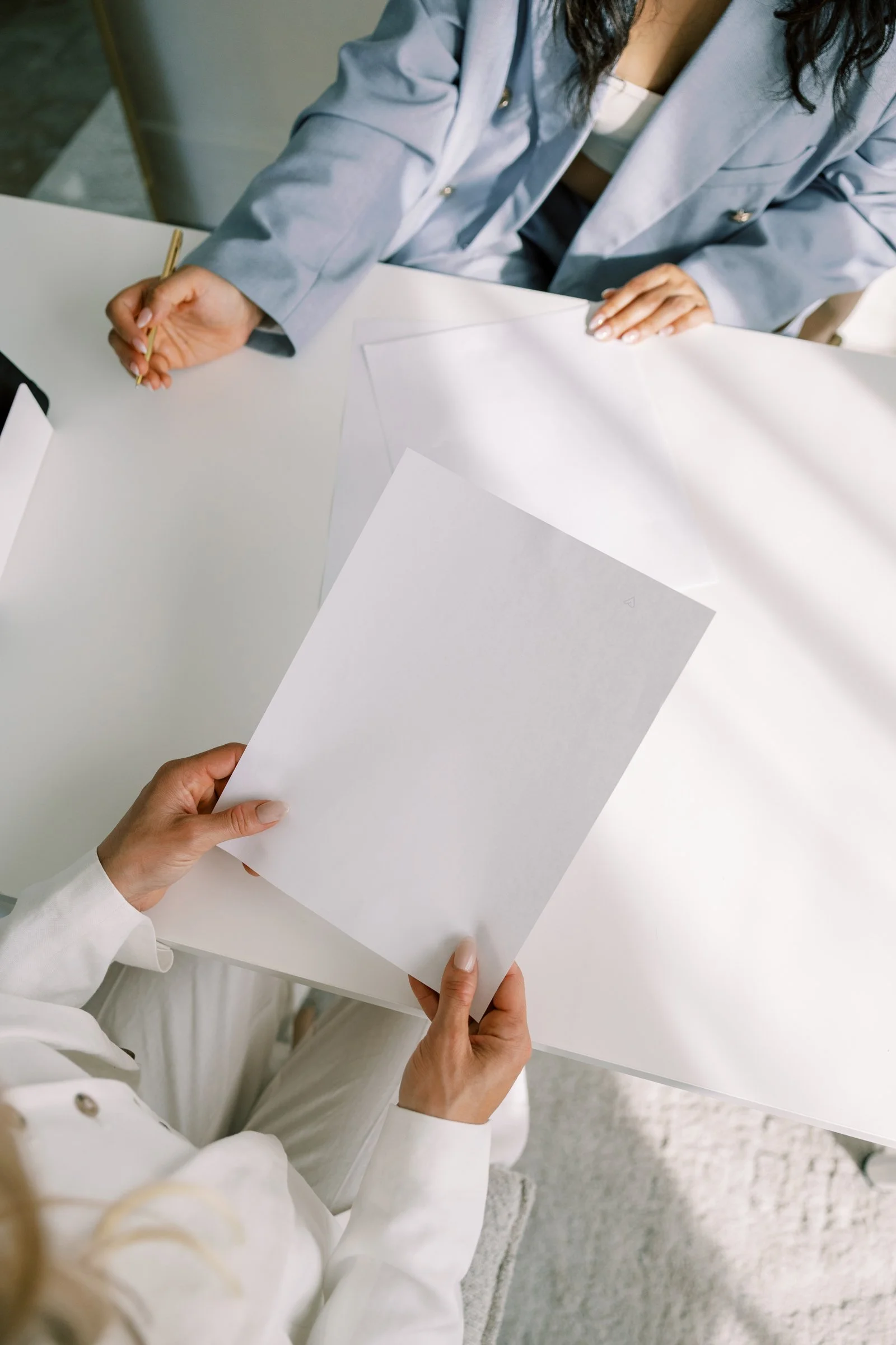 A woman in a light gray blazer sitting at a white table, holding a gold pen, with blank sheets of paper in front of her, and another person's hand holding a blank sheet of paper close to her.