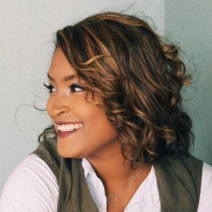 A woman with curly brown hair smiling and looking to her left, wearing a white shirt and a green jacket.
