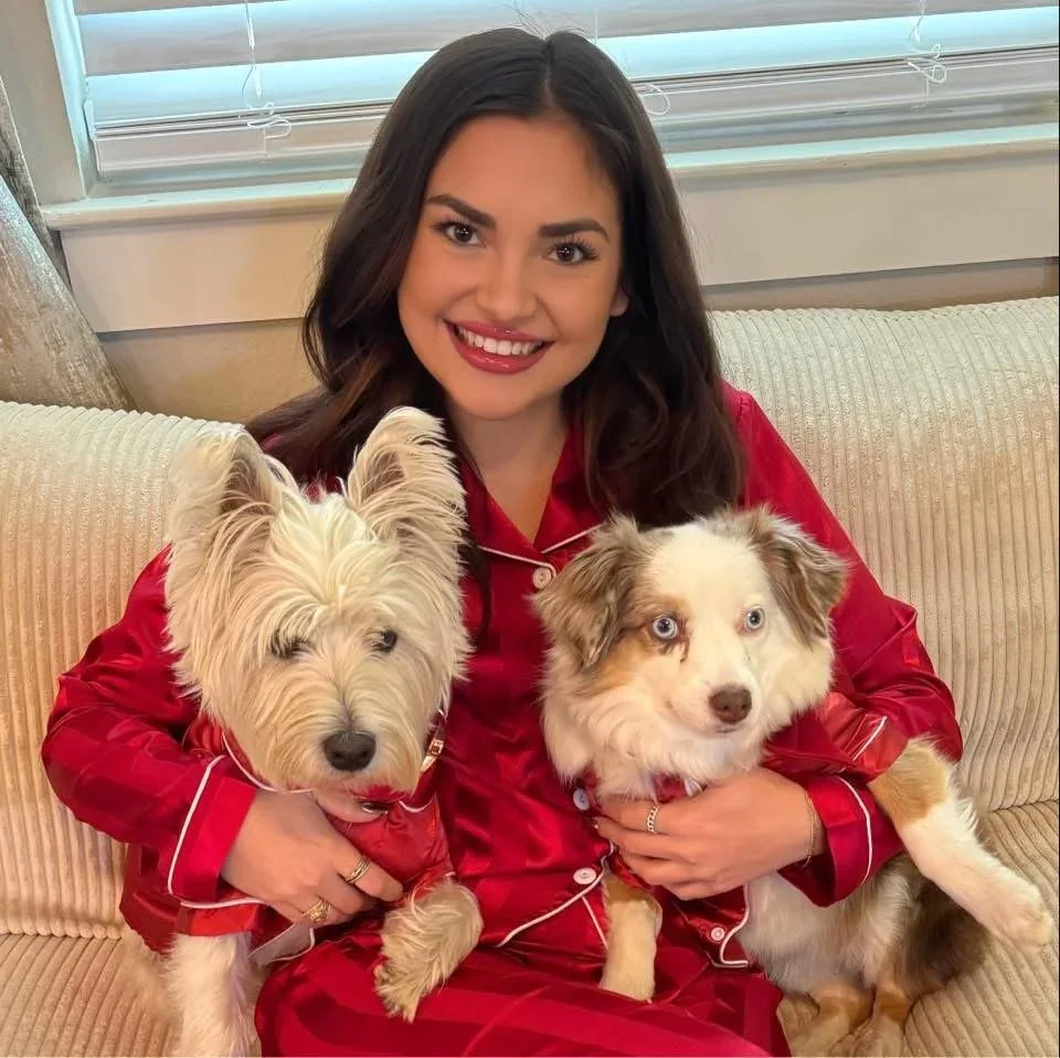 A woman in red pajamas sitting on a beige couch holding two dogs, one white with pointy ears and light fur, the other merle with blue eyes, in front of a window with blinds.