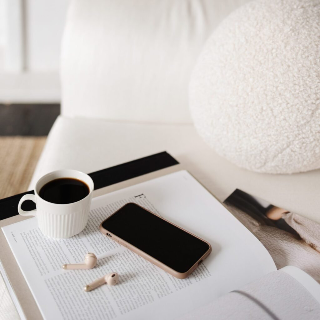 A coffee cup, a smartphone, and wireless earbuds on top of an open book or magazine on a bedside table.