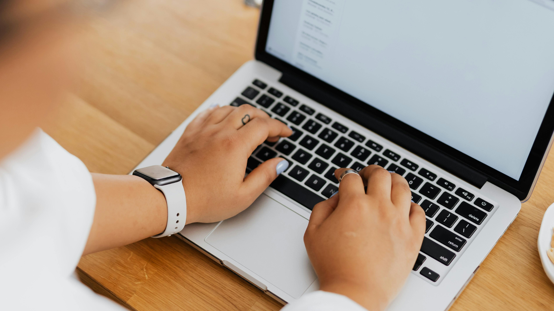 Person using a laptop with a silver body on a wooden desk, wearing a white smartwatch and rings, with a blurred phone in the background.