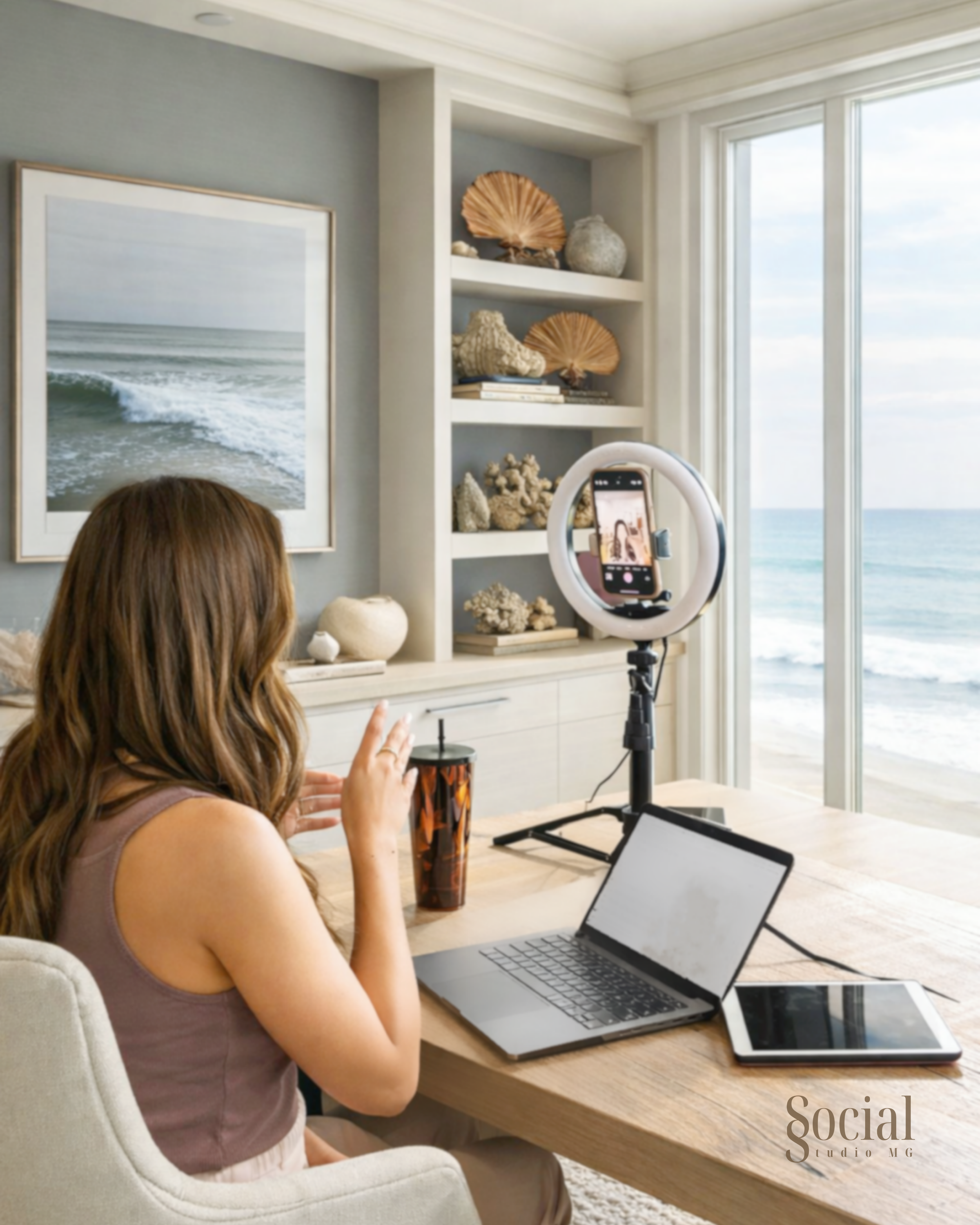 A woman recording herself with a ring light in a coastal home, with a laptop, tablet, and drink on a wooden table, ocean view through large windows.