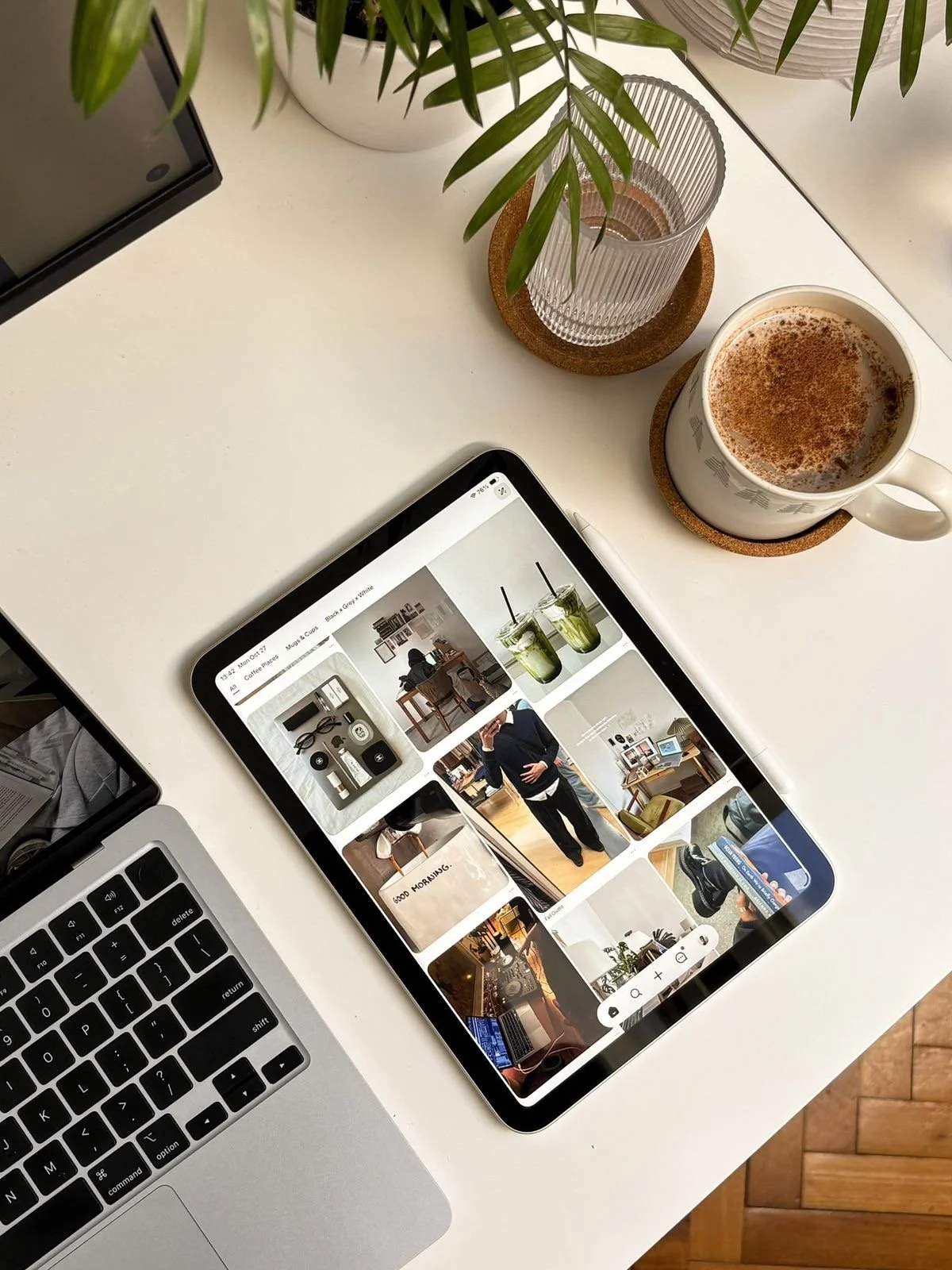 A white desk with a laptop, a tablet displaying a grid of photos, a cup of coffee with froth, a glass of water, and a plant on a white surface.