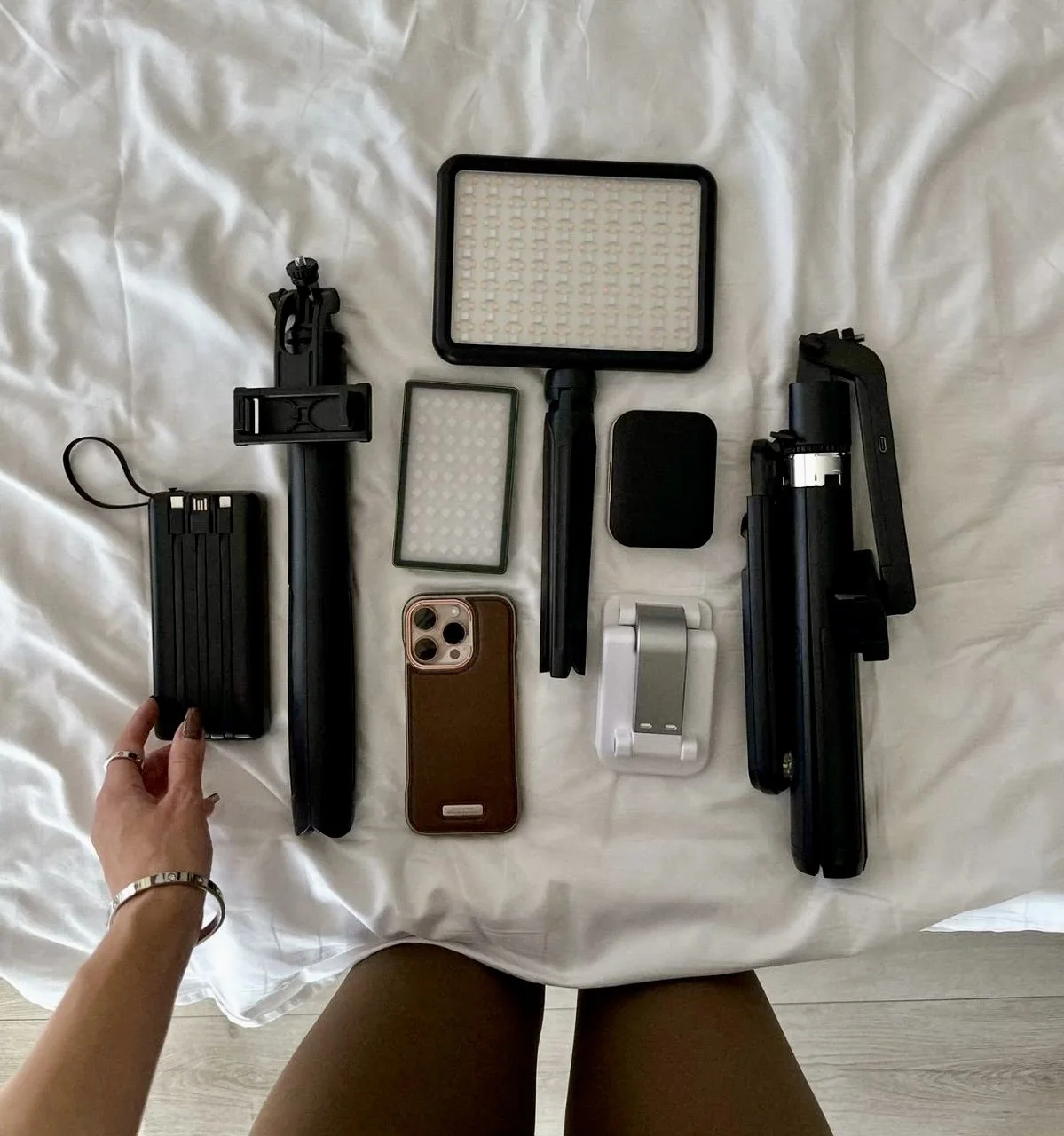 A collection of photography equipment and accessories on a white bedsheet, including a camera tripod, camera filters, a smartphone with a protective case, a small portable device, and various camera parts. A gimbal, and content creation equipment