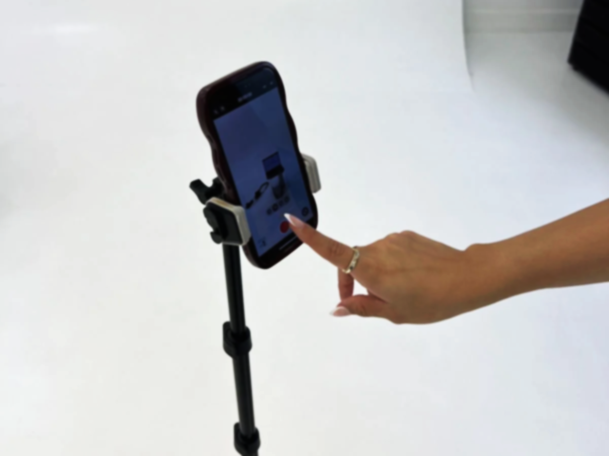 A person's hand using a finger to tap on a smartphone mounted on a selfie stick or tripod stand against a plain white background.