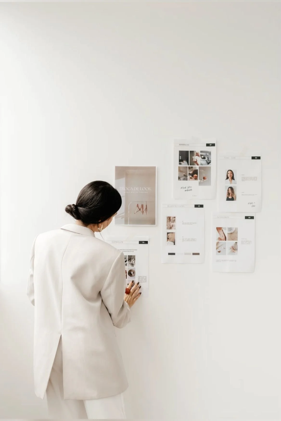 A woman in a white blazer looking at a wall with posters and printed sheets of paper, which include images and text, in a minimalistic setting.