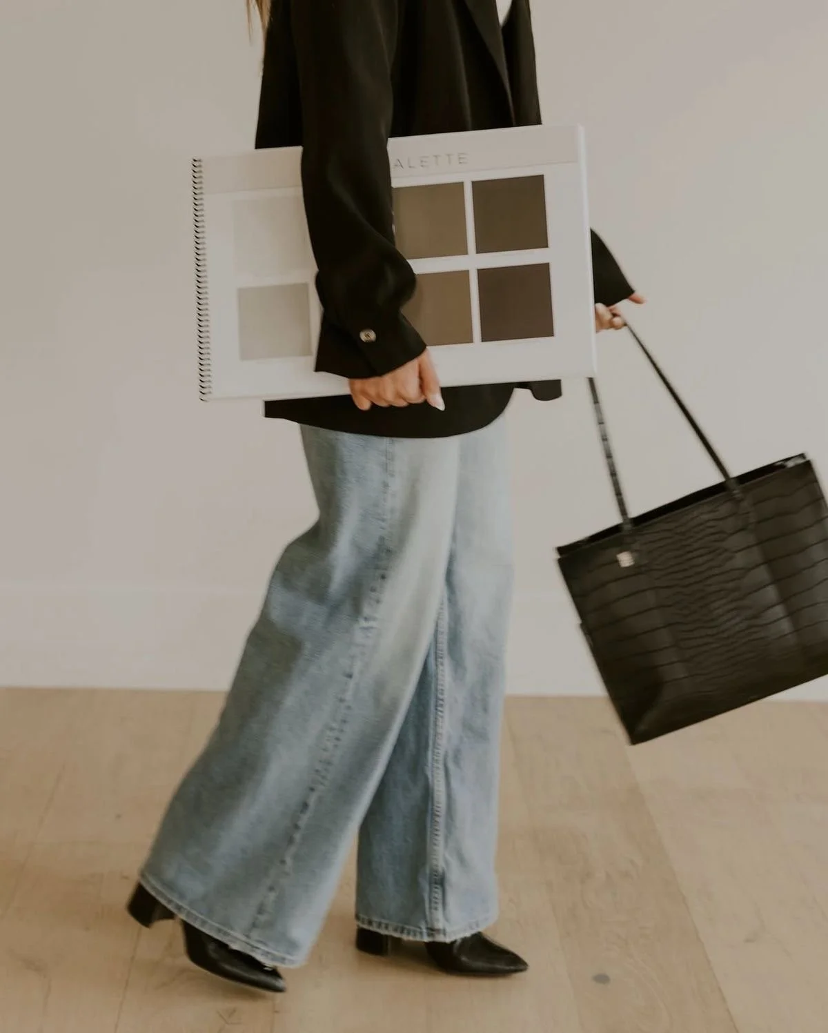 Person wearing a black jacket and wide-leg jeans, holding a color swatch board and a black shopping bag.