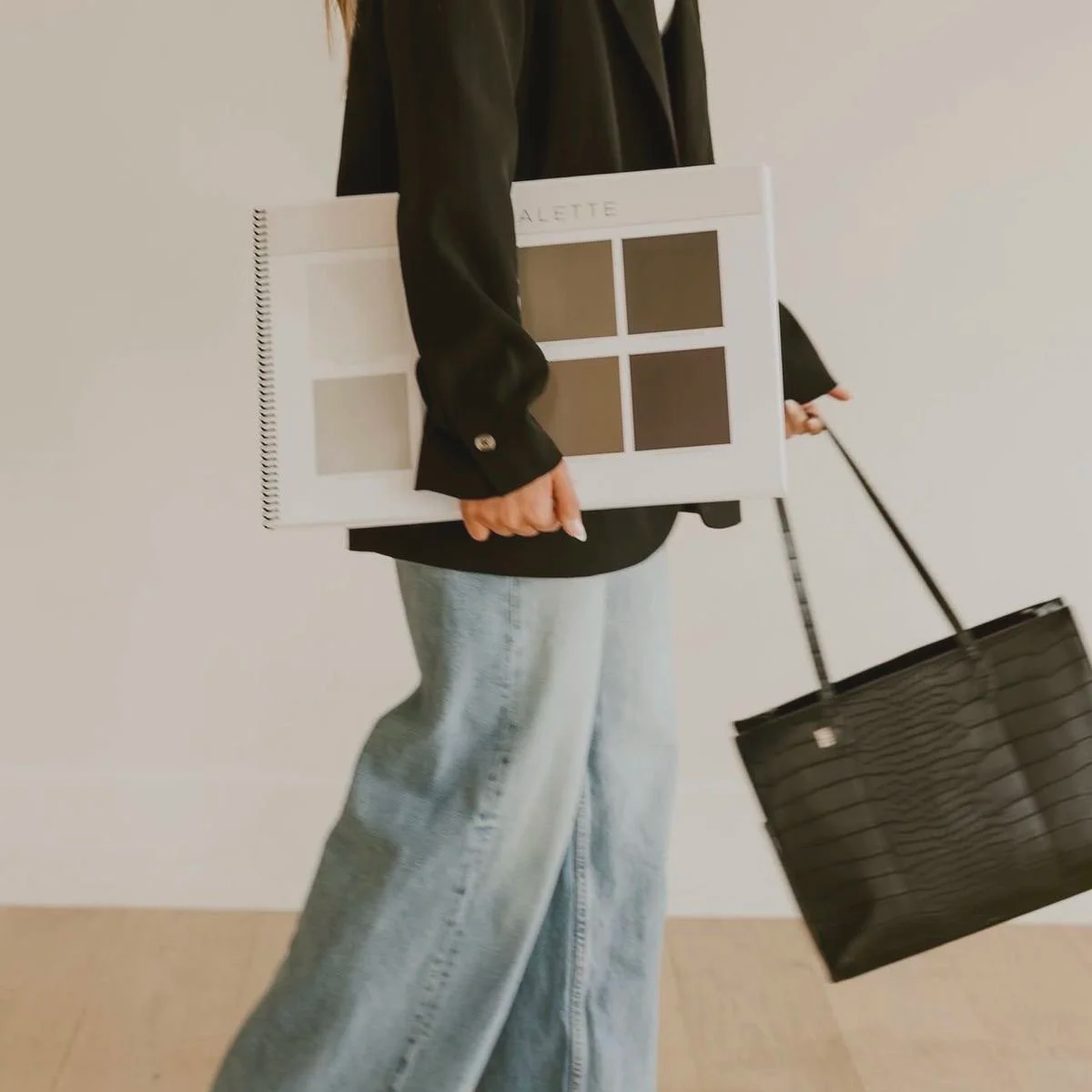 Person holding a color swatch book and a black tote bag while standing against a plain wall. It is a brand design kit