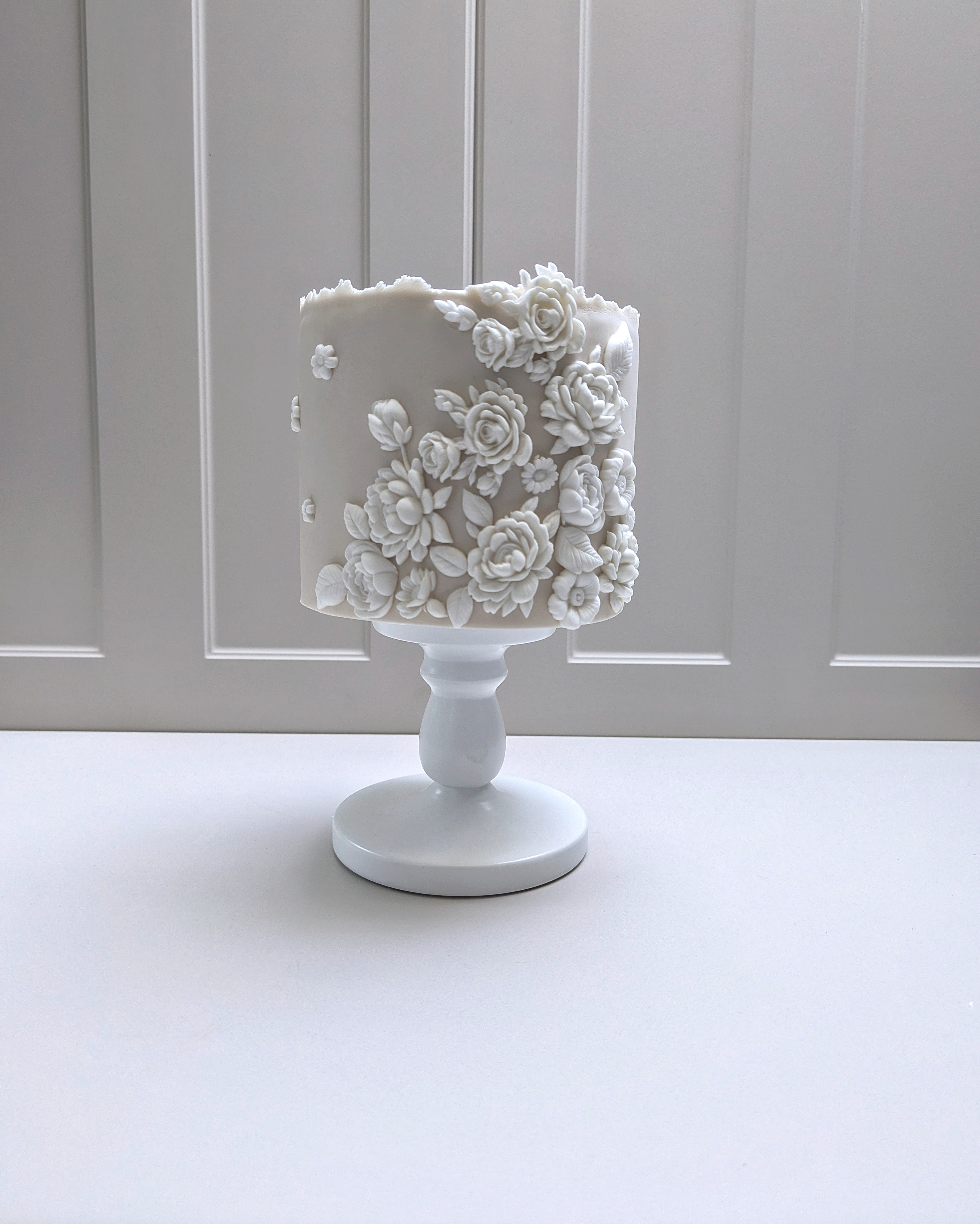 Elegant nude shade fondant covered cake with fondant flower detailing on the side. The cake is displayed on a white cake pedestal.
