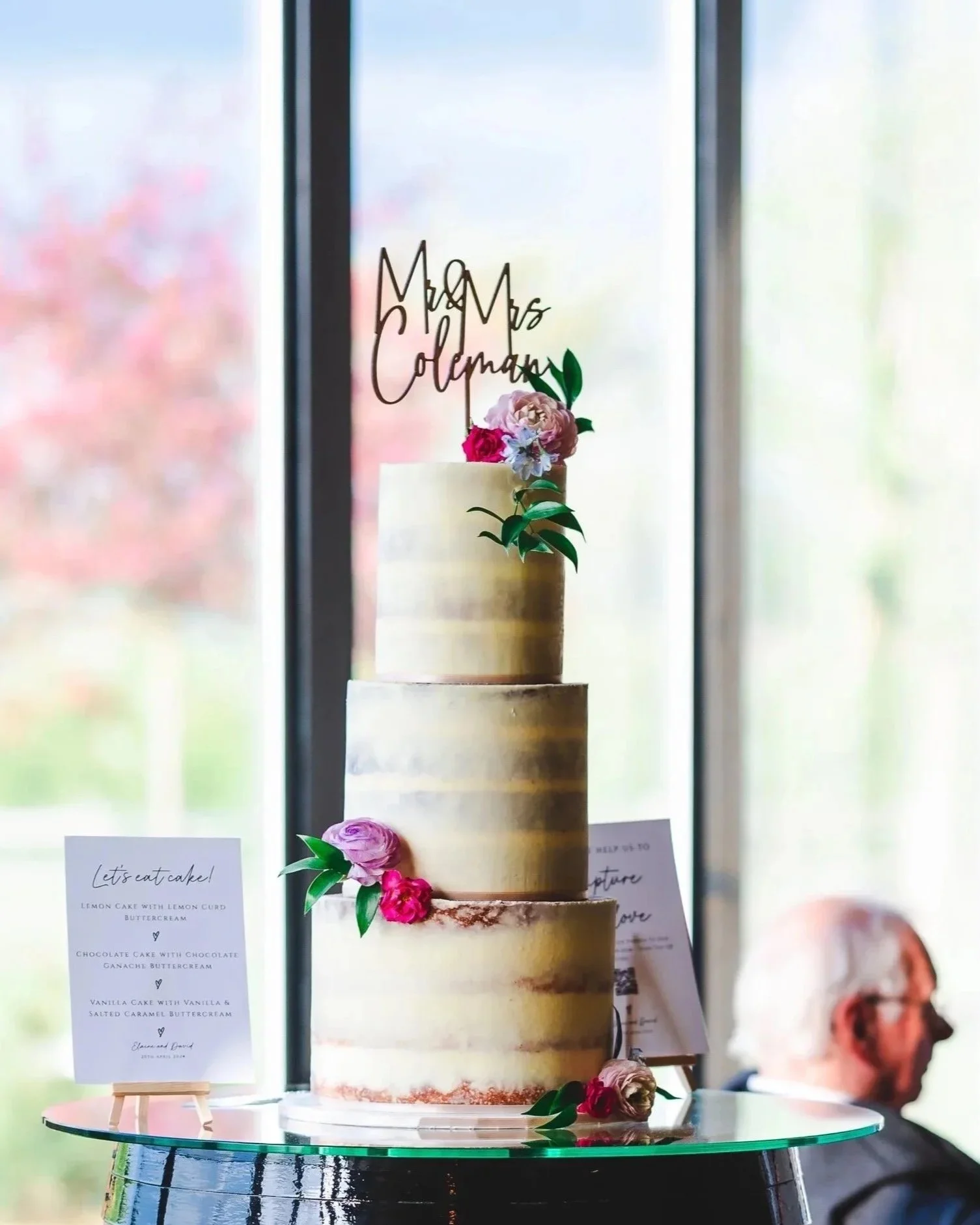 A tall, elegant, 3 tiered semi-naked buttercream style wedding cake decorated with pink and lilac flowers, green leaves, and a wooden cake topper.  The cake is displayed on a glass table in front of a window. Venue: Primrose Hill Farm. Photographer: 
