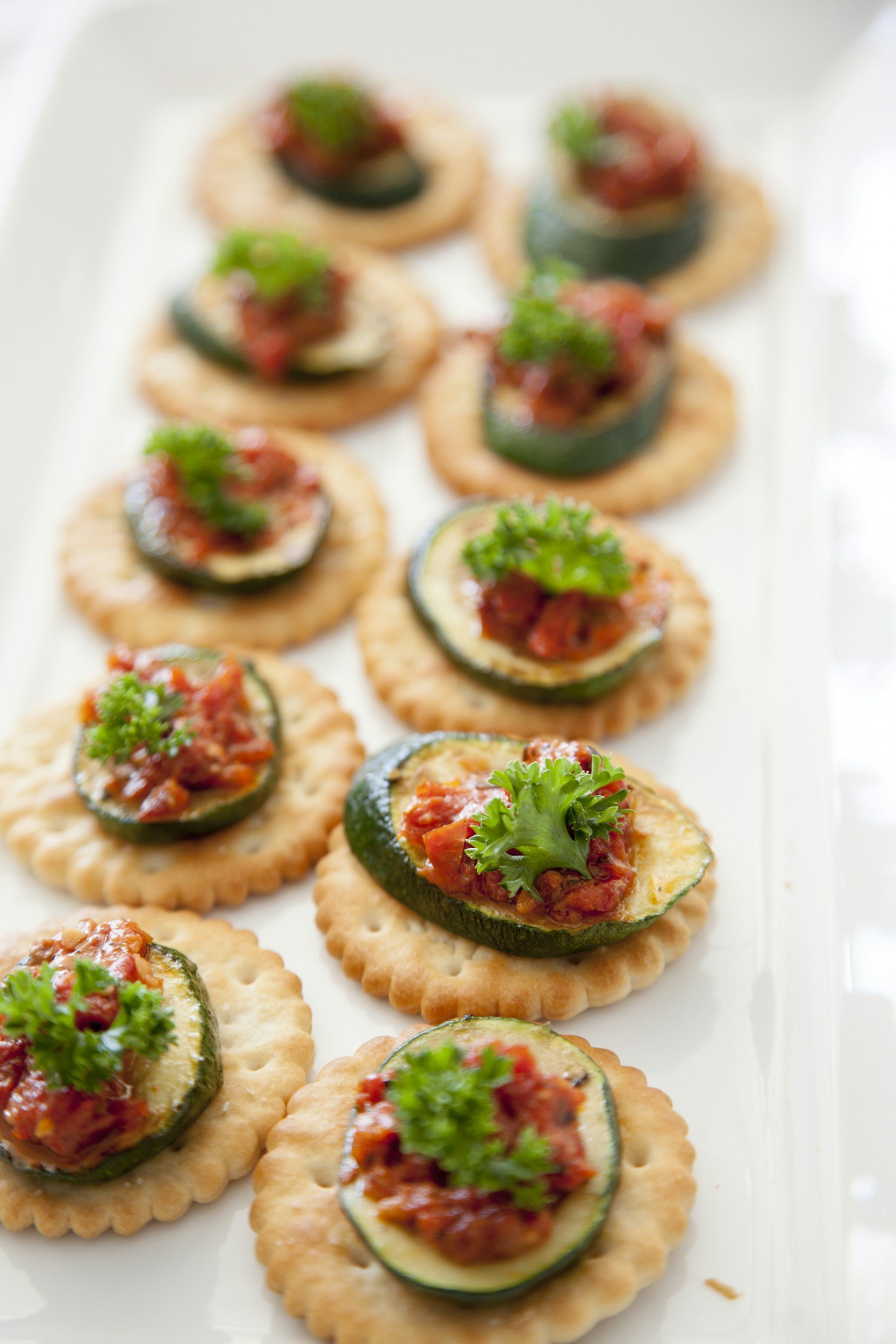 Close-up of round crackers topped with sliced zucchini, a spoonful of tomato-based topping, and a sprig of parsley.