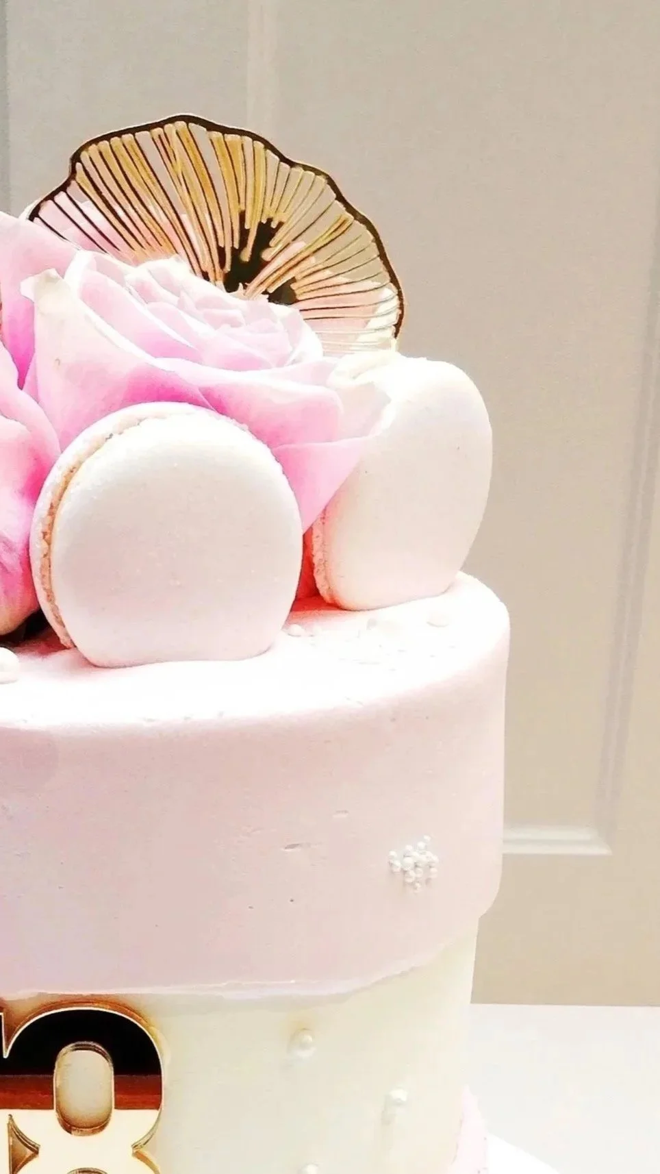 Close-up of a pastel pink birthday cake decorated with pink flowers, white macarons, a golden flower edible decoration, and small white pearls. Elegant, bespoke celebration cake.