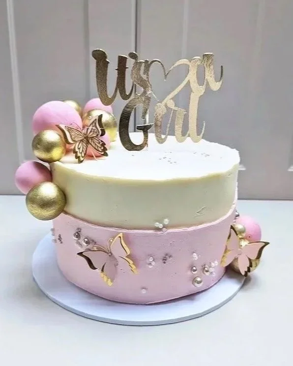 Gorgeous pale pink and ivory buttercream cake with pink and gold spheres, pink and gold butterfly detailing and edible pearls. There is a gold cake topper reading "It's a Girl".
