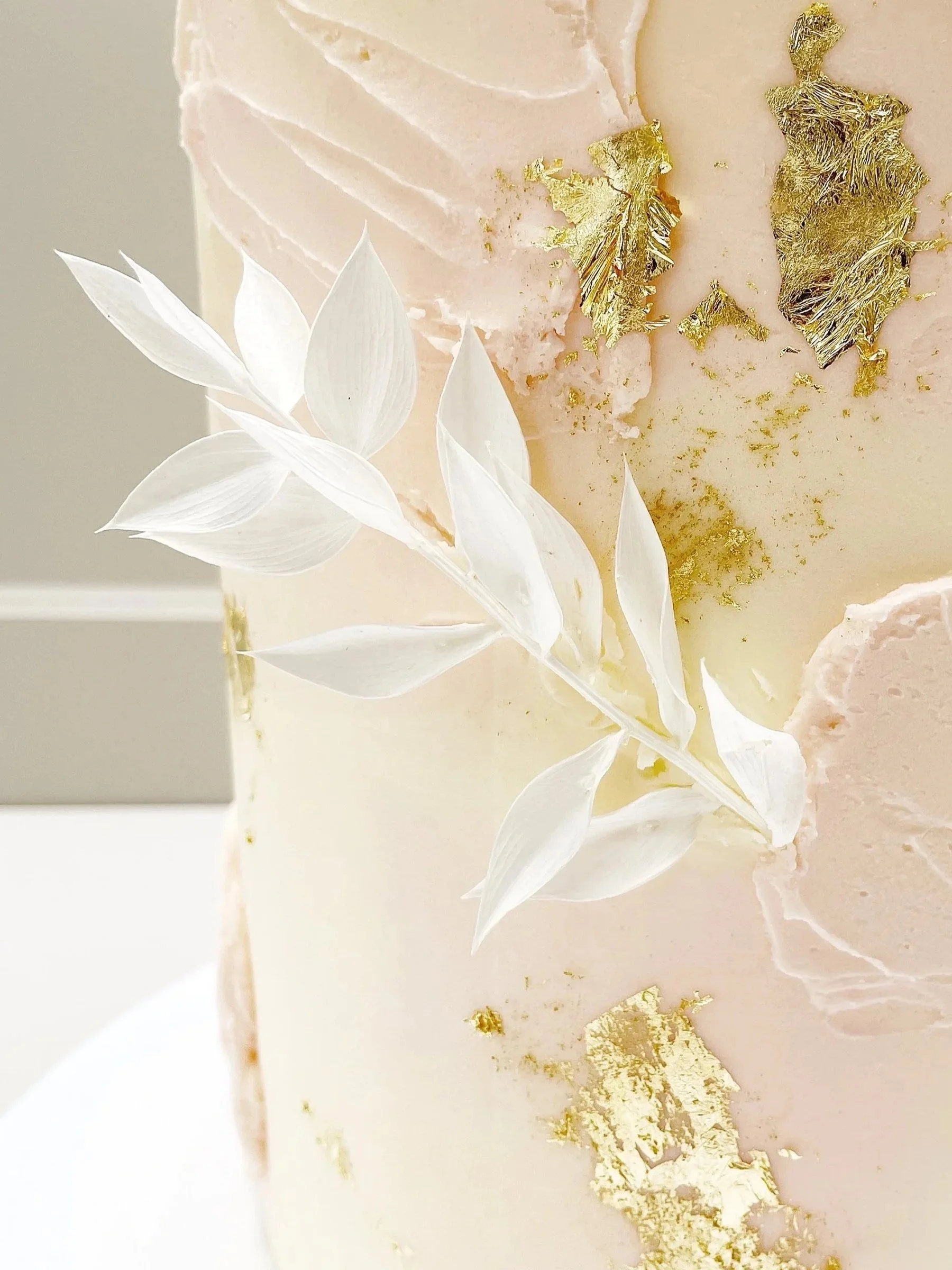 Close-up of a ivory-colored buttercream cake decorated with white leaves, pale pink buttercream texture and gold leaf accents.