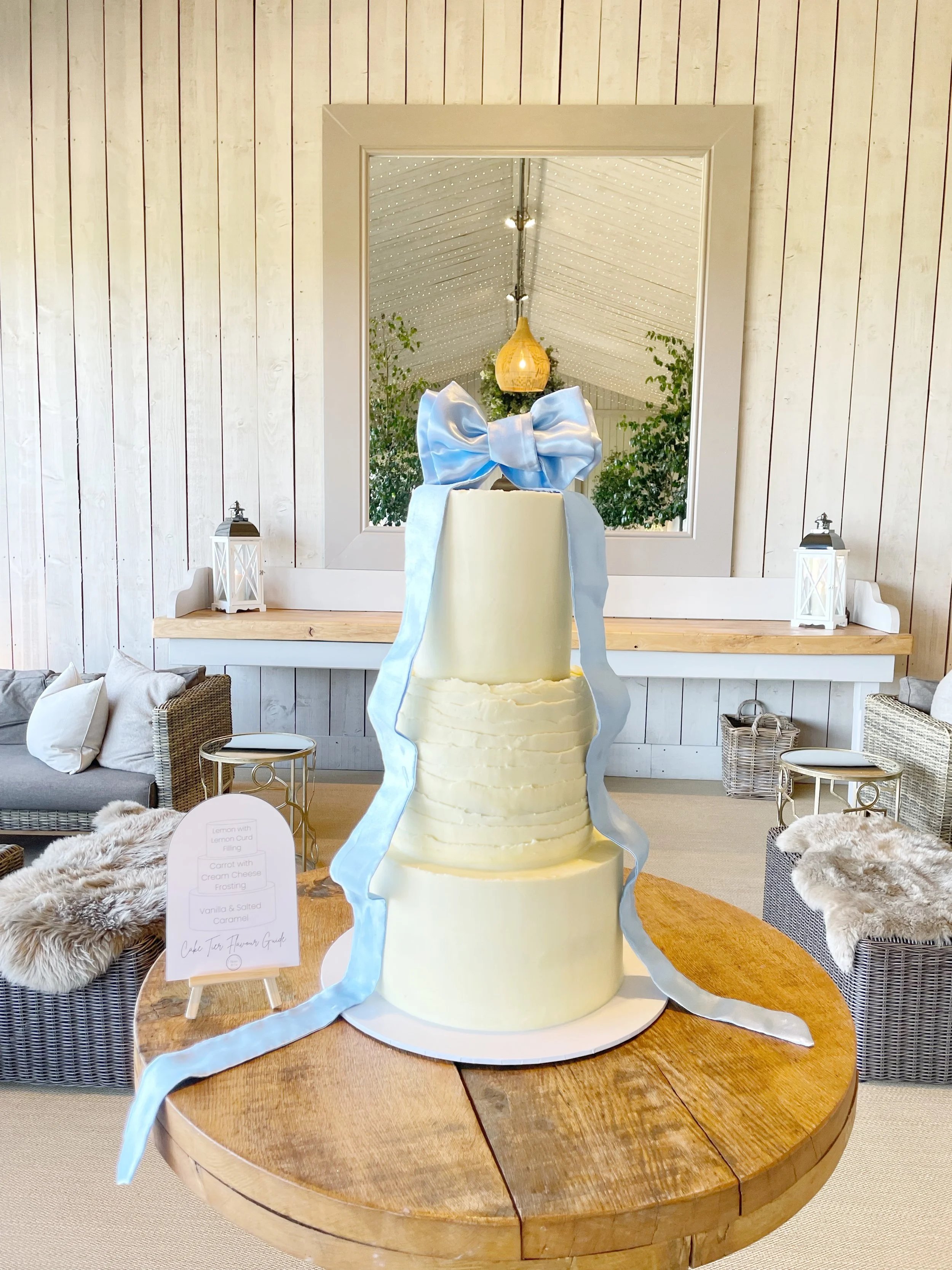 A multi-tiered white cake with a large blue satin bow on top, placed on a wooden table in a cozy, decorated room. The room features a mirror, lanterns, pillows, and a plush rug, with natural wood and white paneling on the walls. Venue: Primrose Hill 