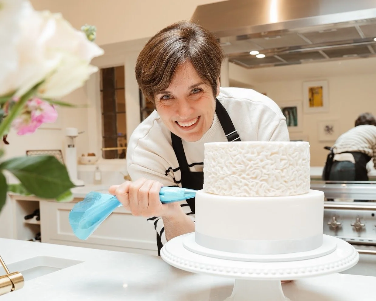 Lisa, owner of Bloon & Co.  decorating a two-tier white cake with textured icing, smiling at the camera in a cozy, well-lit kitchen.