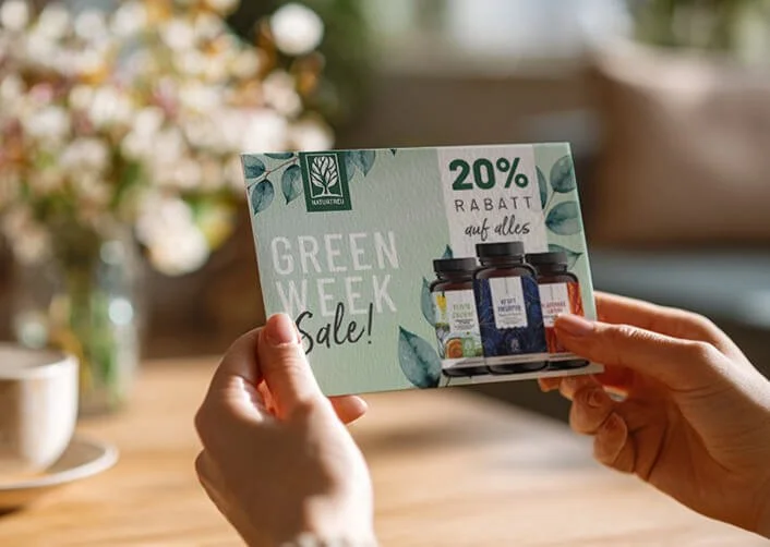 Nahaufnahme einer Hand, die einen Flyer für eine Green Week Sale mit 20 % Rabatt hält, auf einem Holztisch mit Tassen und Pflanzen im Hintergrund.