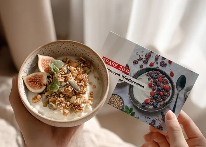 Eine Hand hält eine Schale mit Joghurt, garniert mit Feigen, Granola und Kernen. In der anderen Hand wird eine Werbeflyer über Müslischüssel mit Beeren gehalten.
