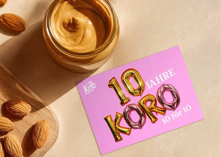 Eine Tafel mit Nutella, eine Karte mit der Aufschrift '10 Jahre KORO, 10 für 10', einige Mandeln auf einem Holzbrett.