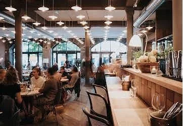 Interior of a lively cafe with patrons seated at tables and a bar counter, large windows, and modern decor.