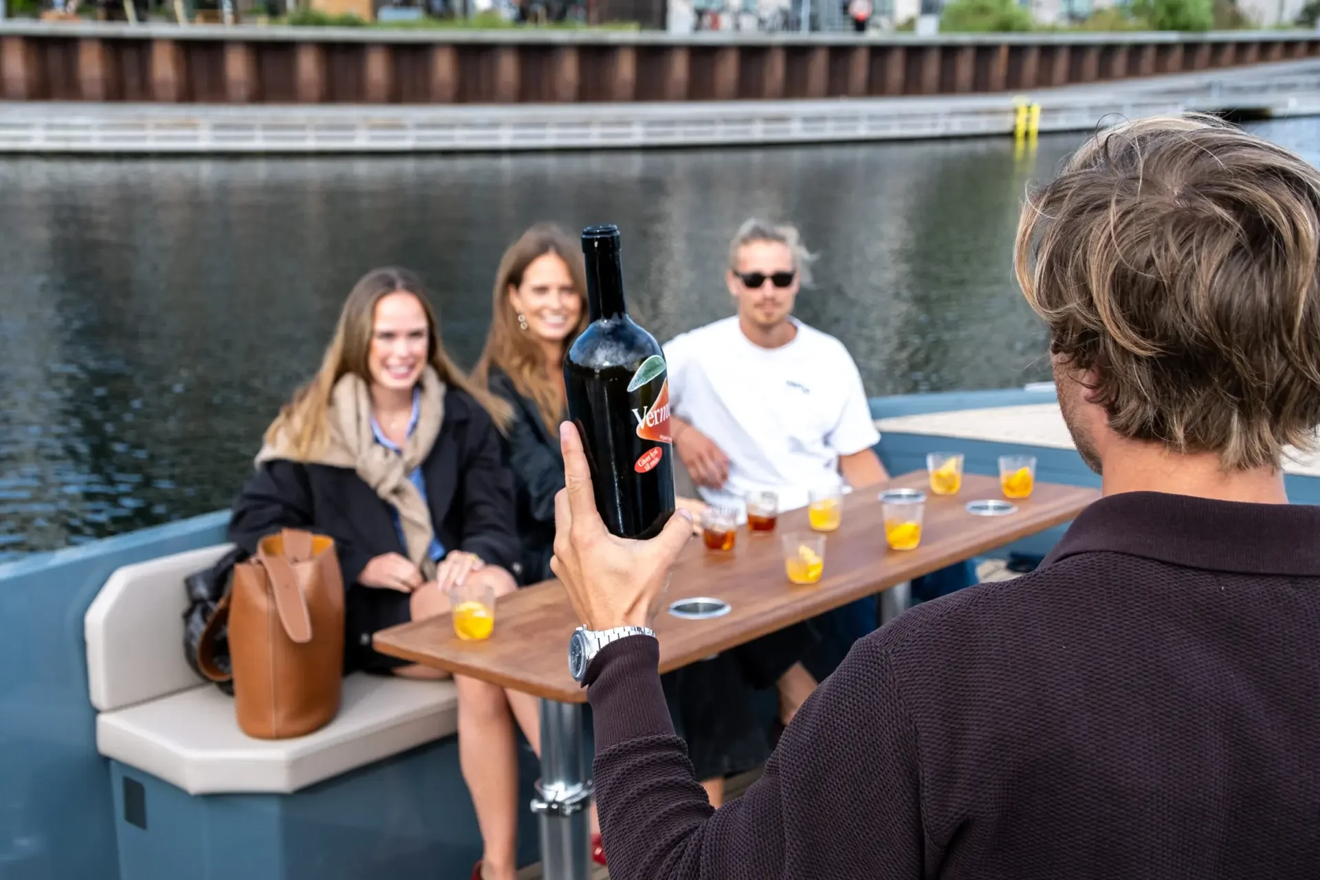 A person is holding a bottle of red wine on a boat with three women seated at a table by the water. The women are smiling and enjoying drinks, and the background shows water and a shoreline with trees.