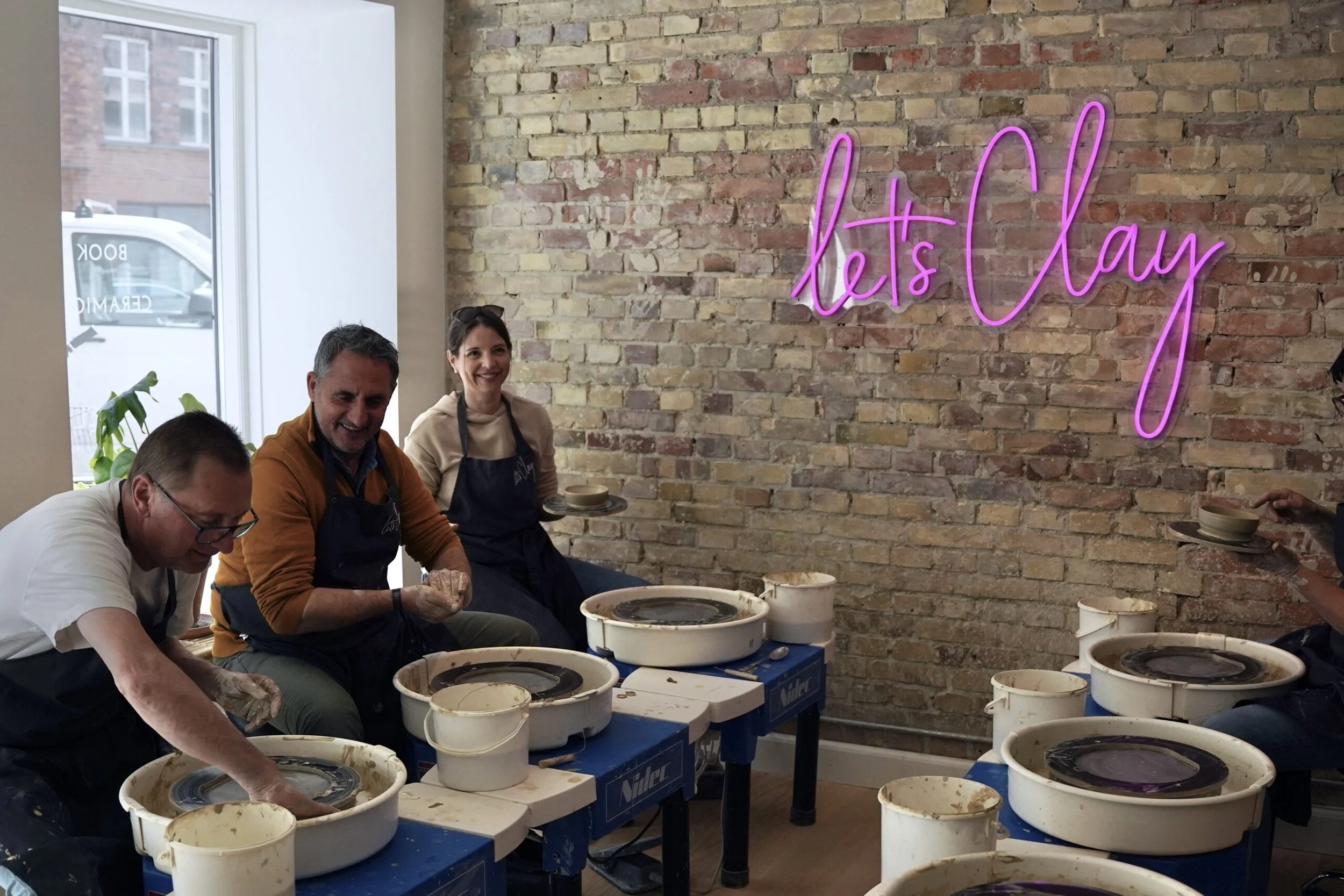 Four people participating in a pottery class, using pottery wheels, in a room with exposed brick wall and a neon sign that reads 'let's Clay' in pink, during daytime.