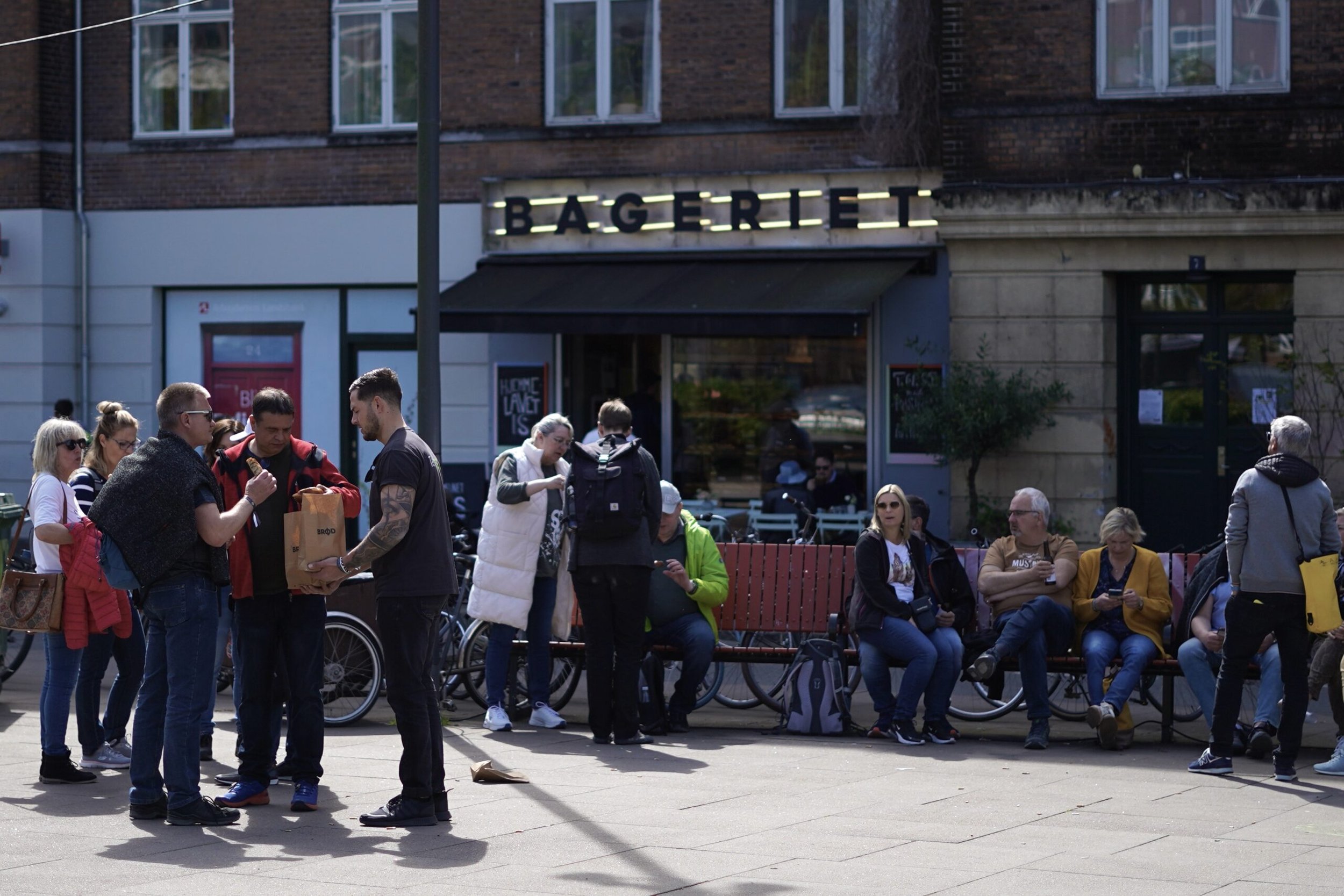 People sitting on benches and standing outside a bakery, some looking at their phones and others talking, in front of a building with the sign 'BAGERIET'.