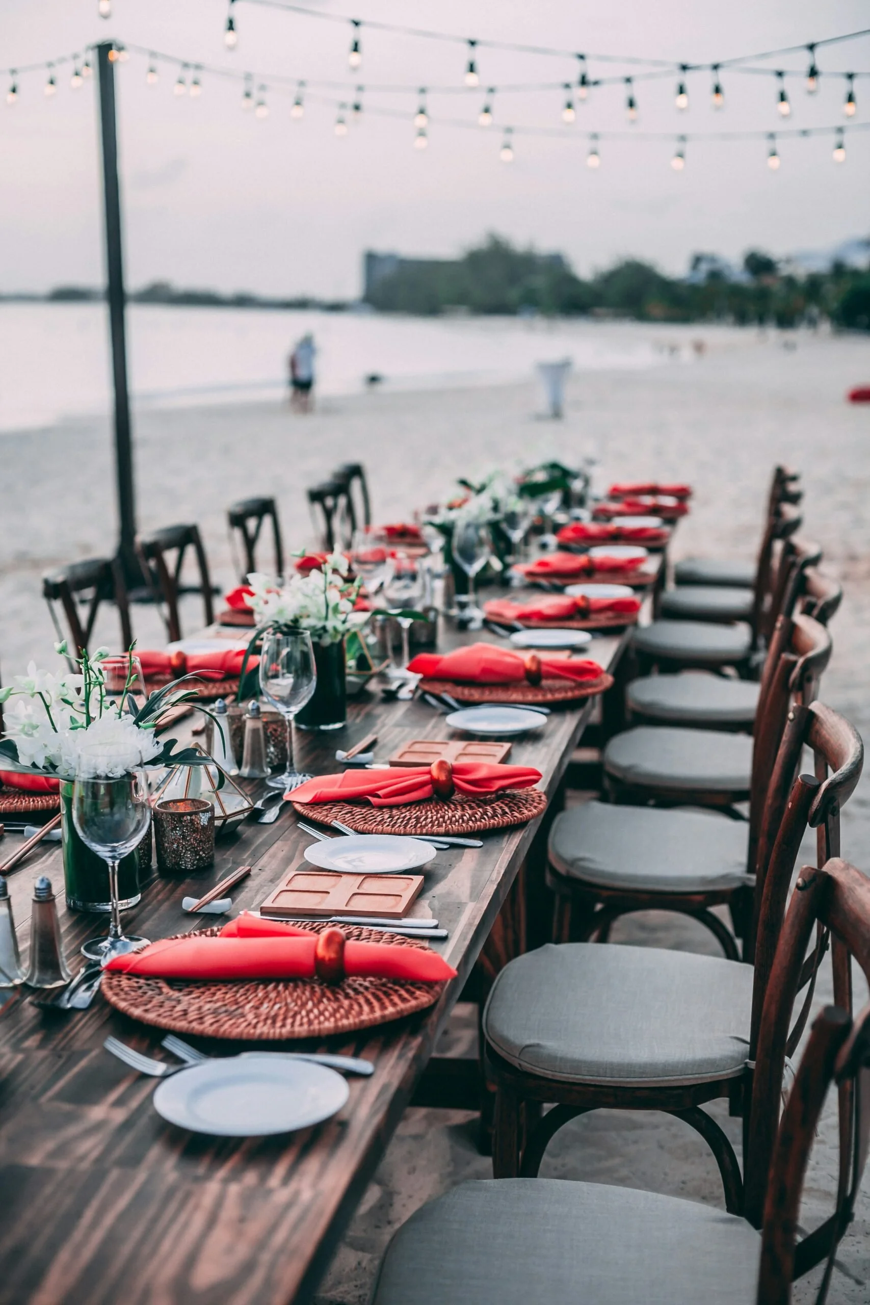 Elegantly set outdoor dining table on the beach with string lights overhead, arranged with red napkins, wine glasses, white plates, and floral centerpieces.