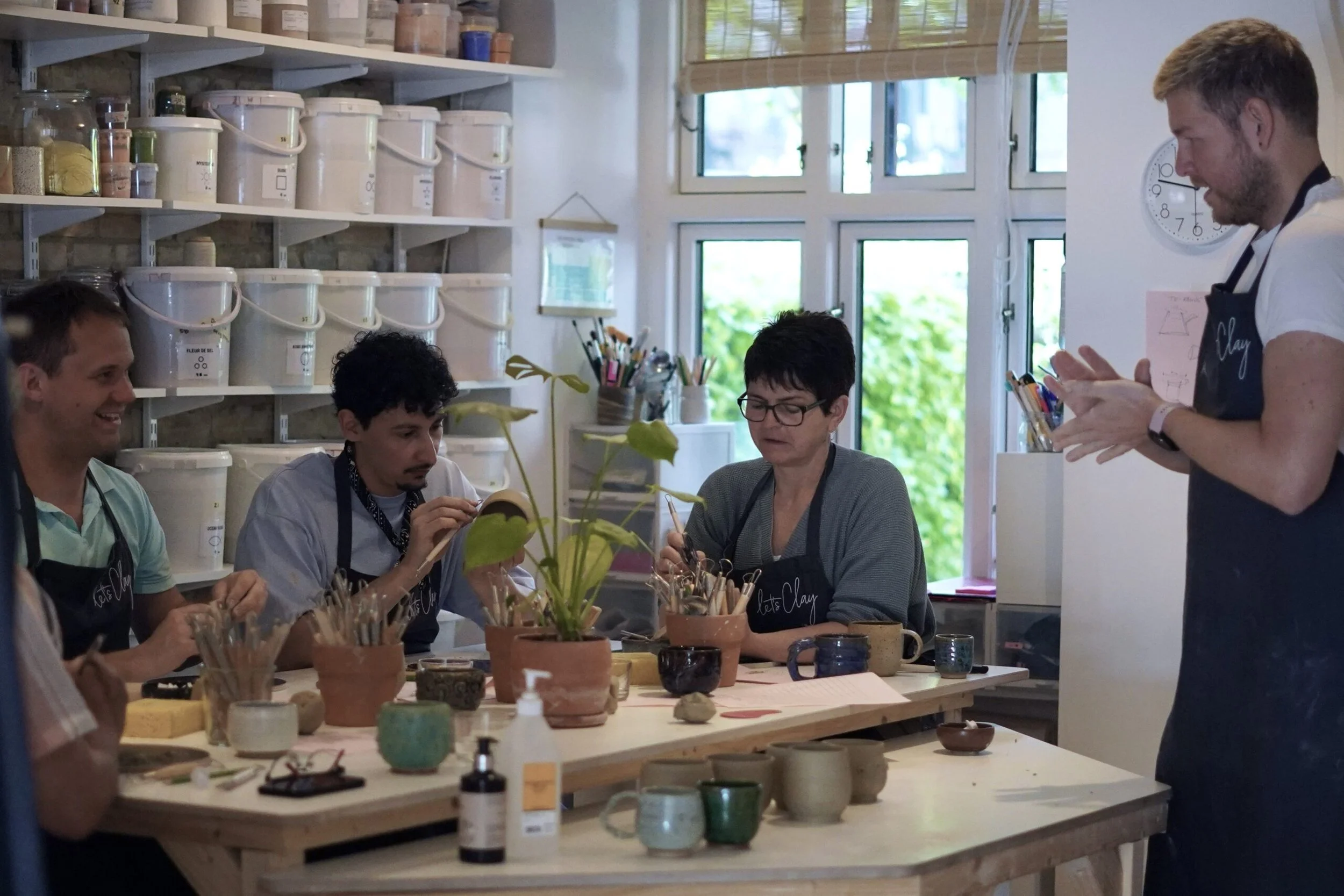 People participating in a pottery crafting class in a well-lit studio with shelves of supplies, working on clay projects around a table.