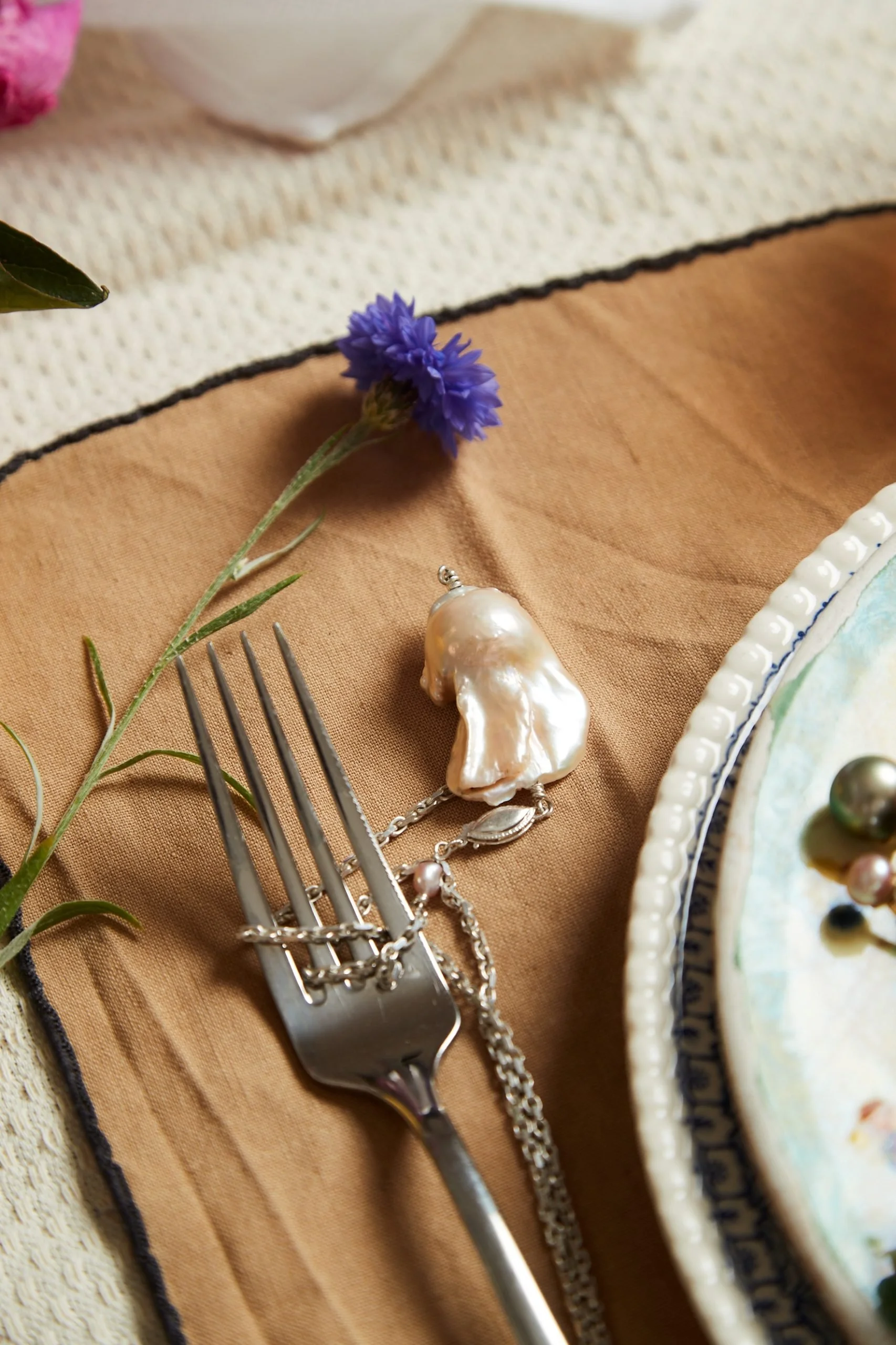 A silver dinner fork with jewelry and a pearl pendant, a purple flower, and ceramic plates with decorative pearls on a beige and brown tablecloth.