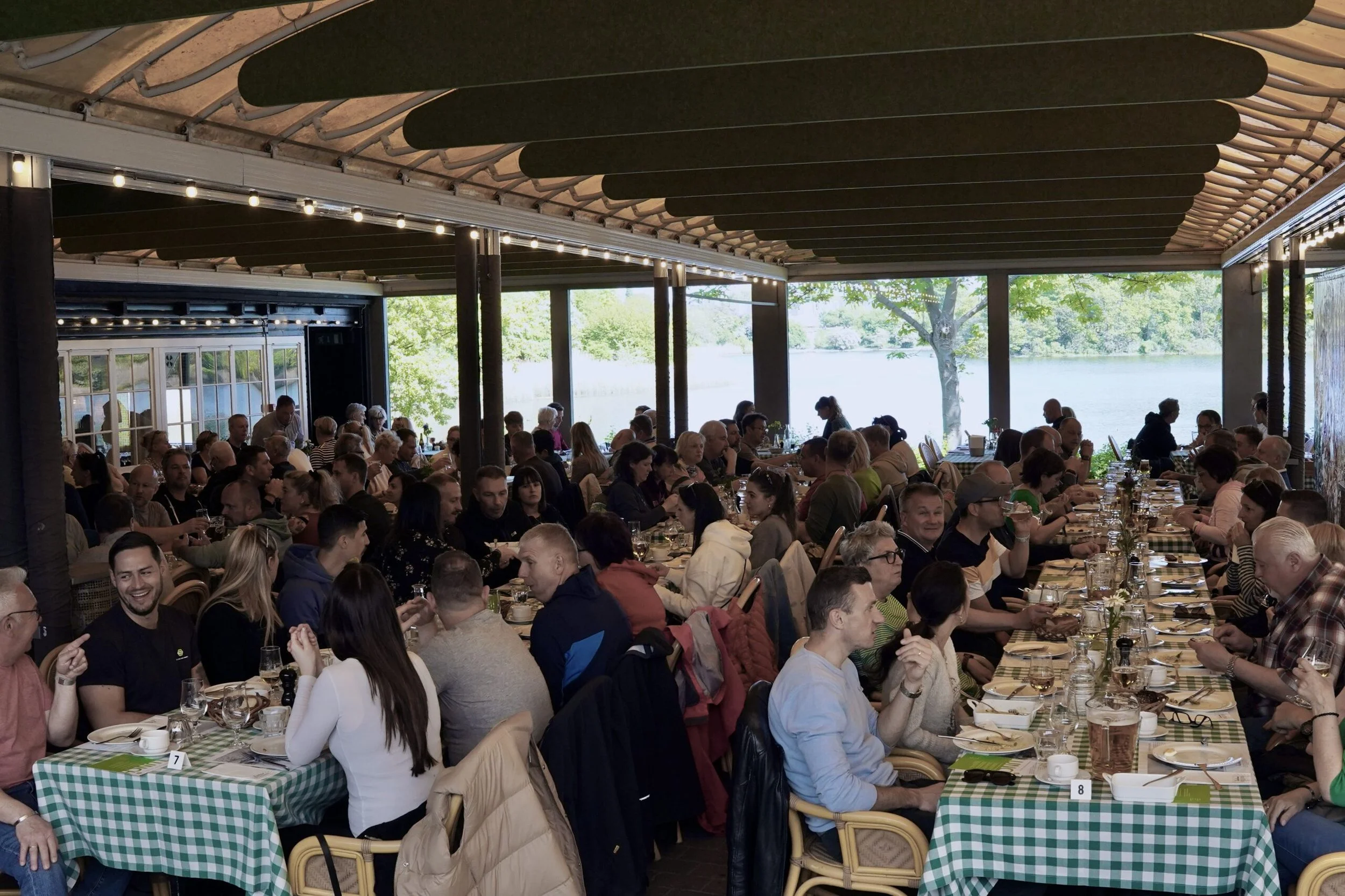 Large group of people dining at long tables with green checkered tablecloths, enjoying a meal in a bright restaurant with large windows overlooking a lake and trees.