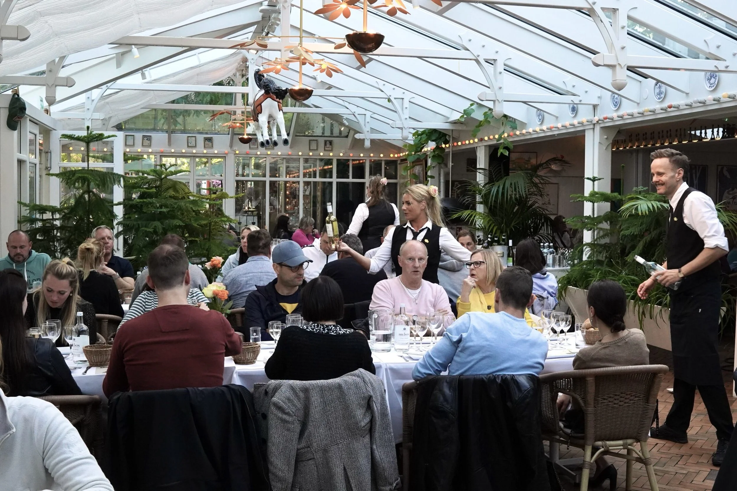 People dining in a glass-roofed restaurant with plants and a carousel horse decoration hanging from the ceiling, with waitstaff serving wine and drinks.