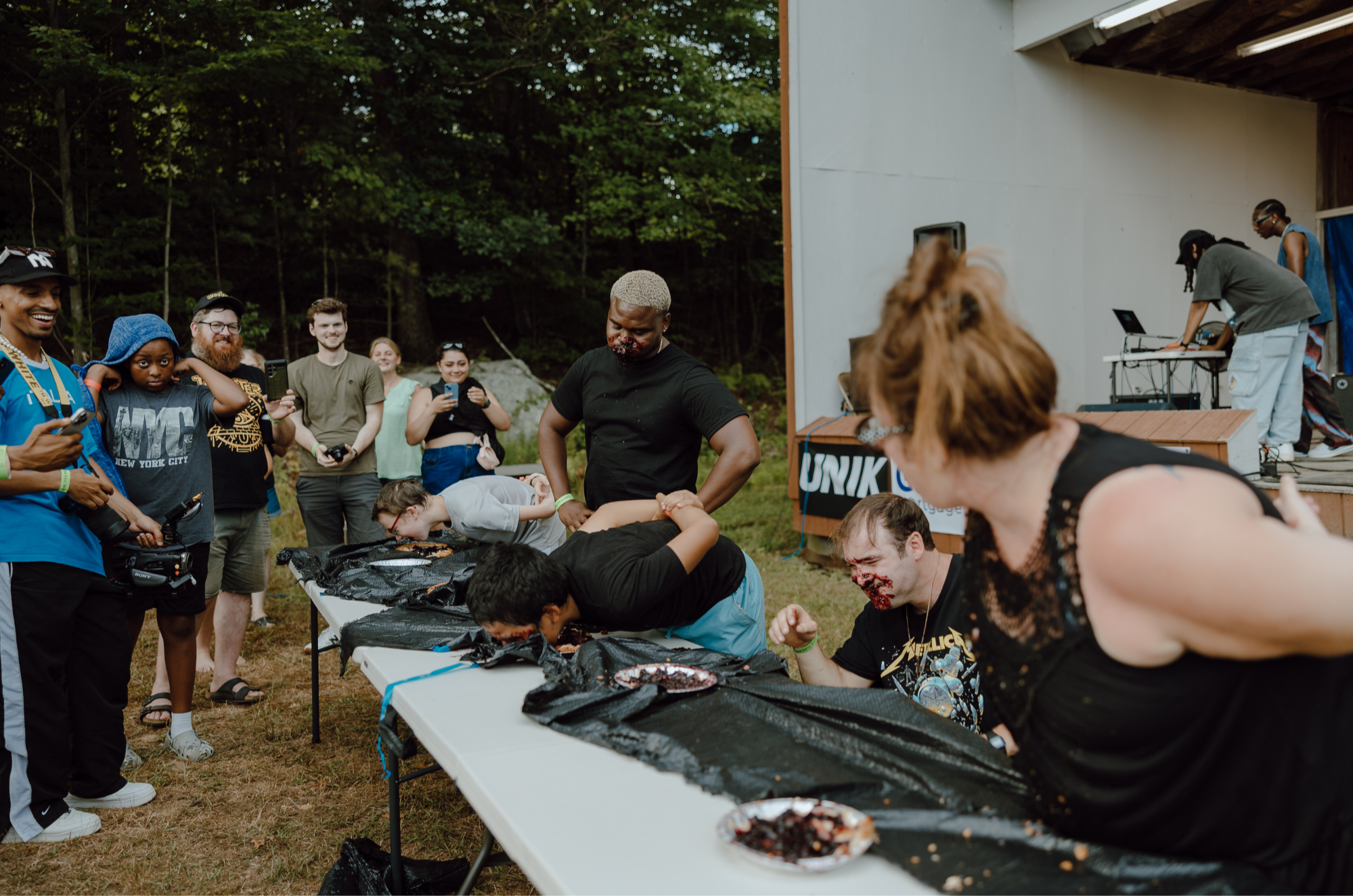 People participating in a pie-eating contest outdoors, with crowd watching and a DJ on a stage in the background.