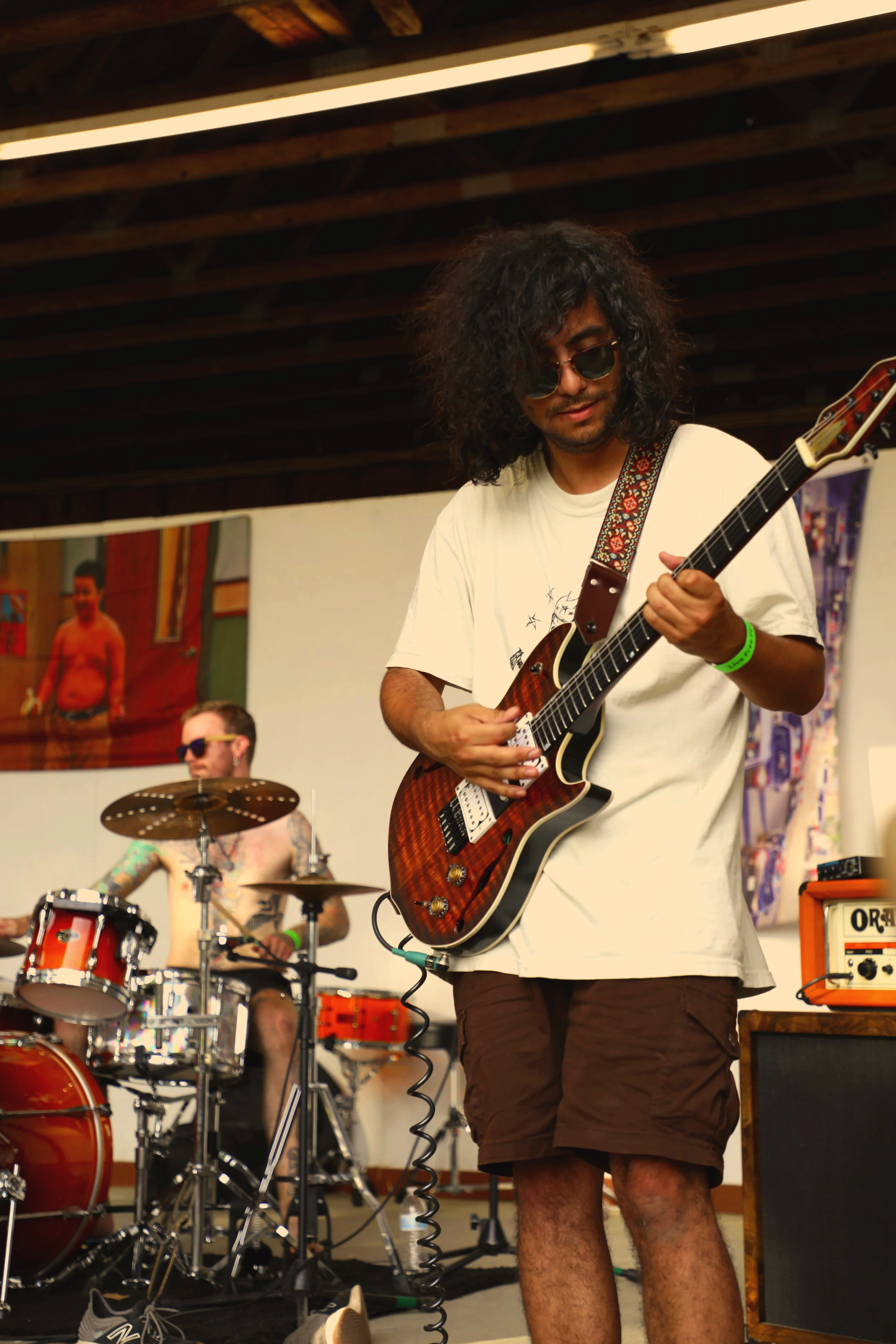 A musician with curly hair and sunglasses playing an electric guitar, and a drummer with tattoos playing drums in the background during a live band performance.