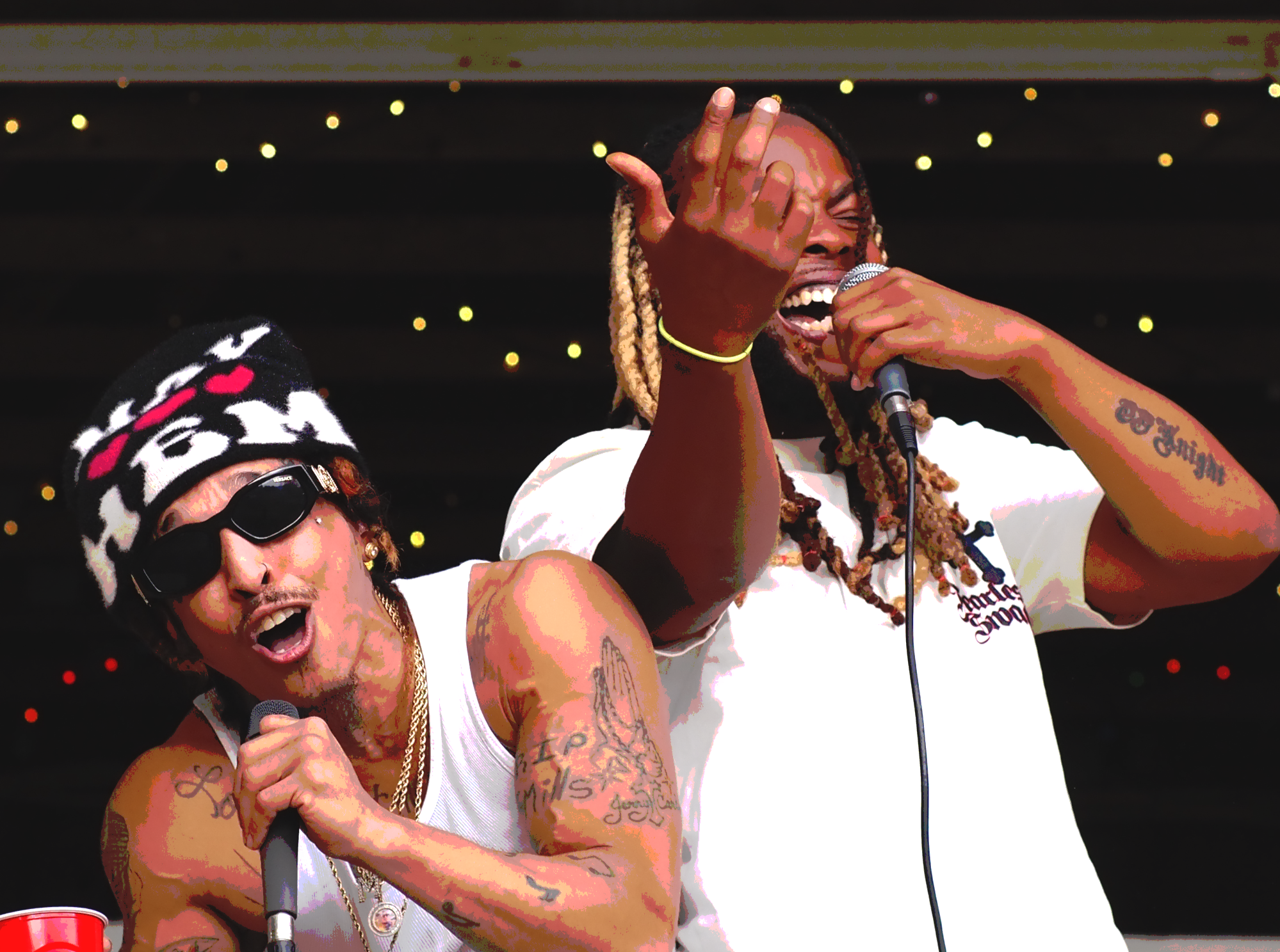 Two musicians singing passionately on stage, one wearing sunglasses and a beanie, the other with dreadlocks and tattoos, both holding microphones, with a dark background.