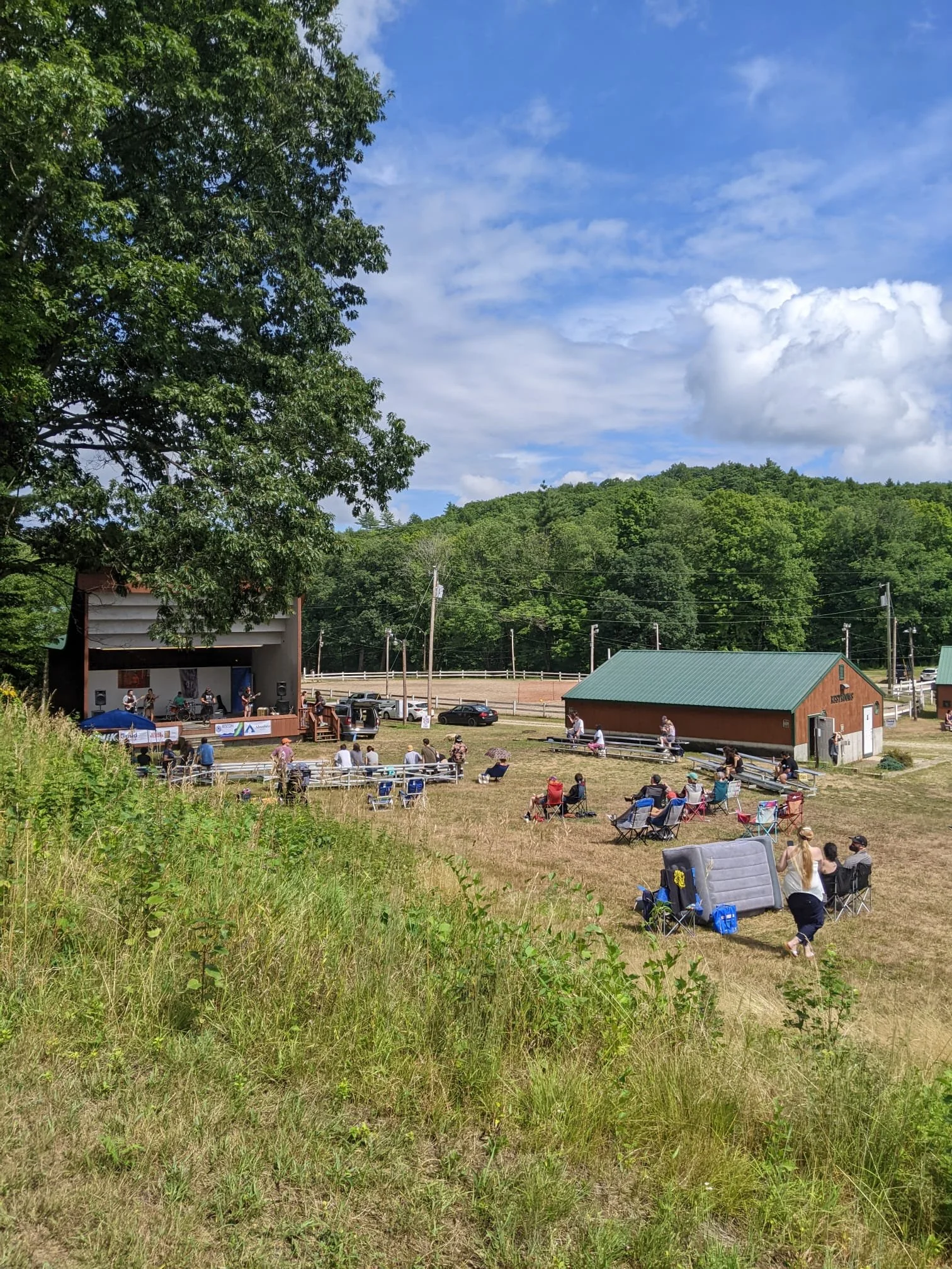 Outdoor concert or event in a grassy field with a small stage, audience seated on chairs and blankets, in a rural area with trees and a hill in the background.