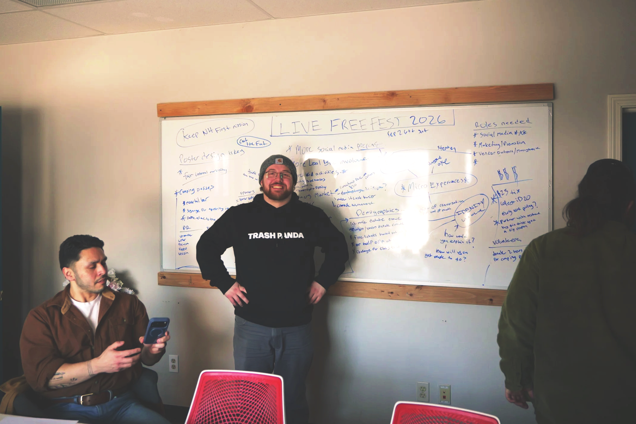 A group of three people in a meeting room with a whiteboard. One person is sitting and looking at a phone, the other two are standing near the whiteboard which has notes and diagrams. The person standing in the center is smiling and wearing a black hoodie that says 'TRASH PANDA'.