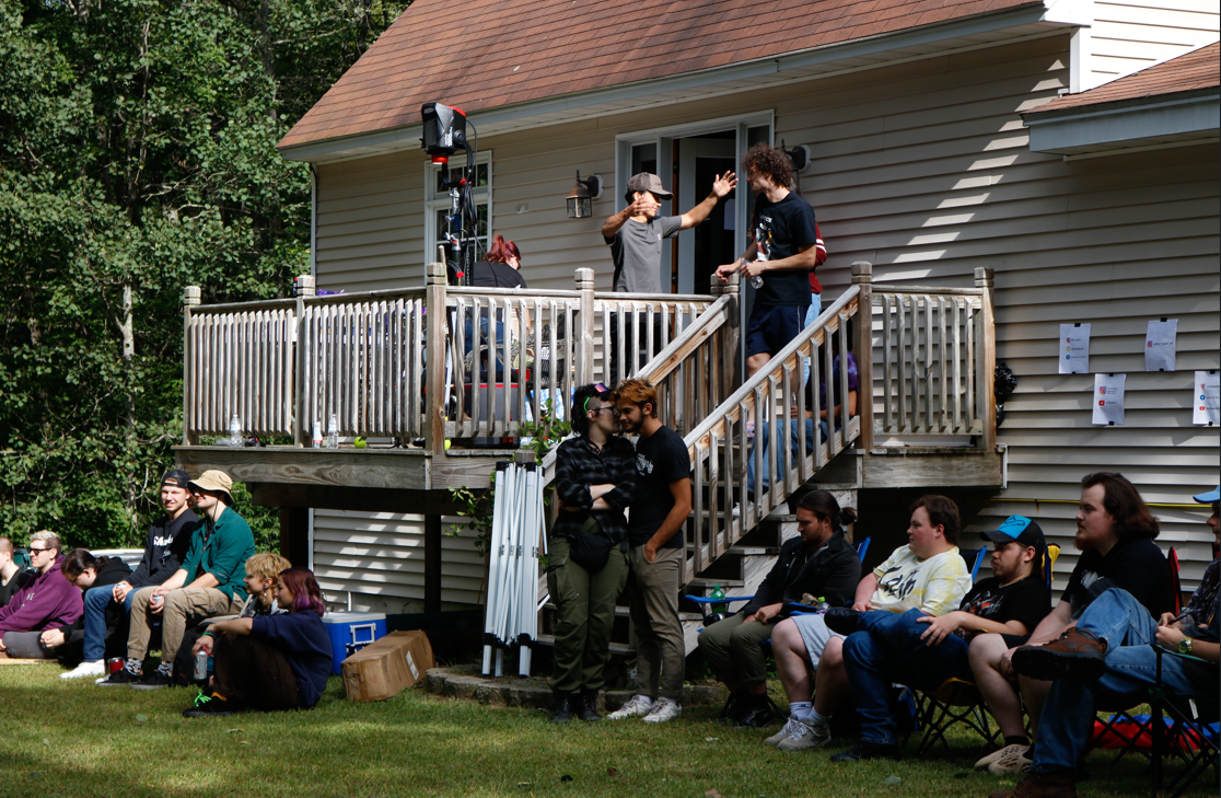 People gathered outdoors for a social event or concert with a backyard setting, some sitting on the grass and others standing or on a deck, with a house and trees in the background.