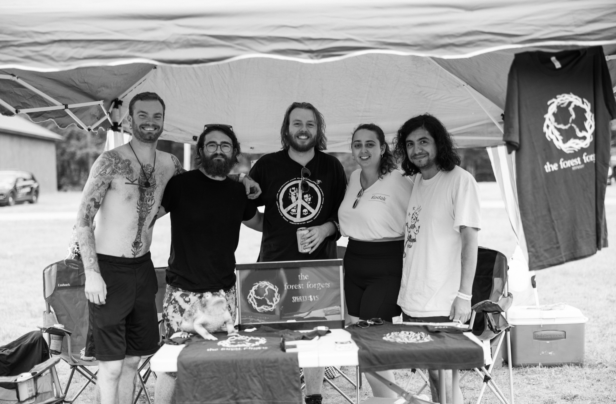 A group of five people standing under a canopy at an outdoor event, smiling at the camera. Their table displays T-shirts and merchandise with a logo of a circle of trees and the text "the forest forgets." One person has tattoos, and another is holding a drink.