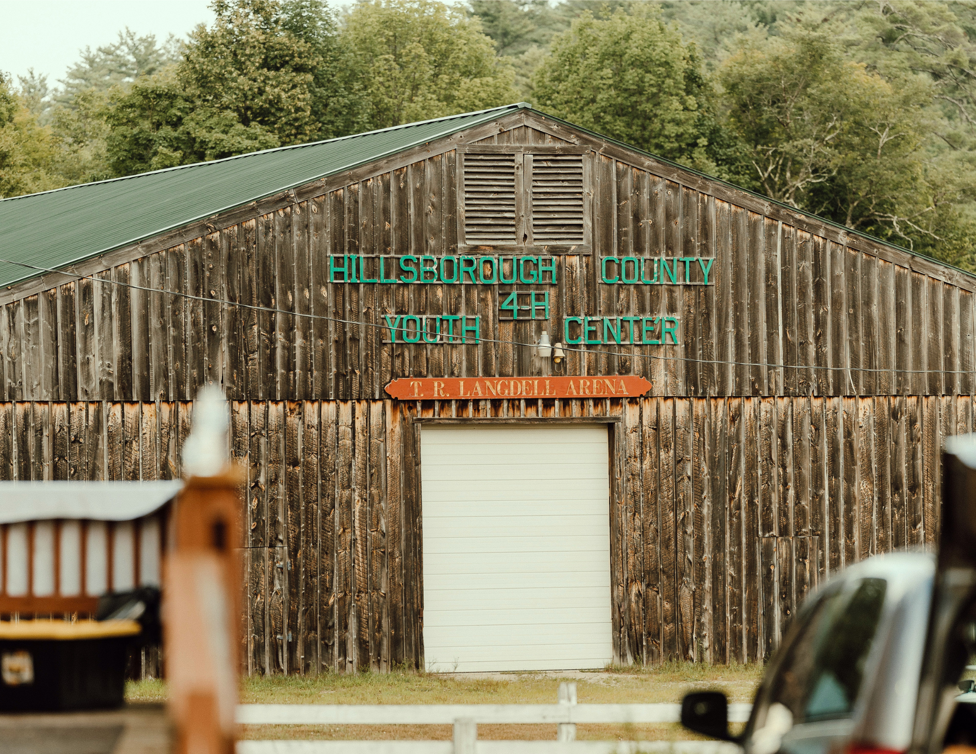 Wooden building with green roof and signs reading 'Hillsborough County 4-H Youth Center' and 'T. R. Langdell Arena' in front of a wooded area.