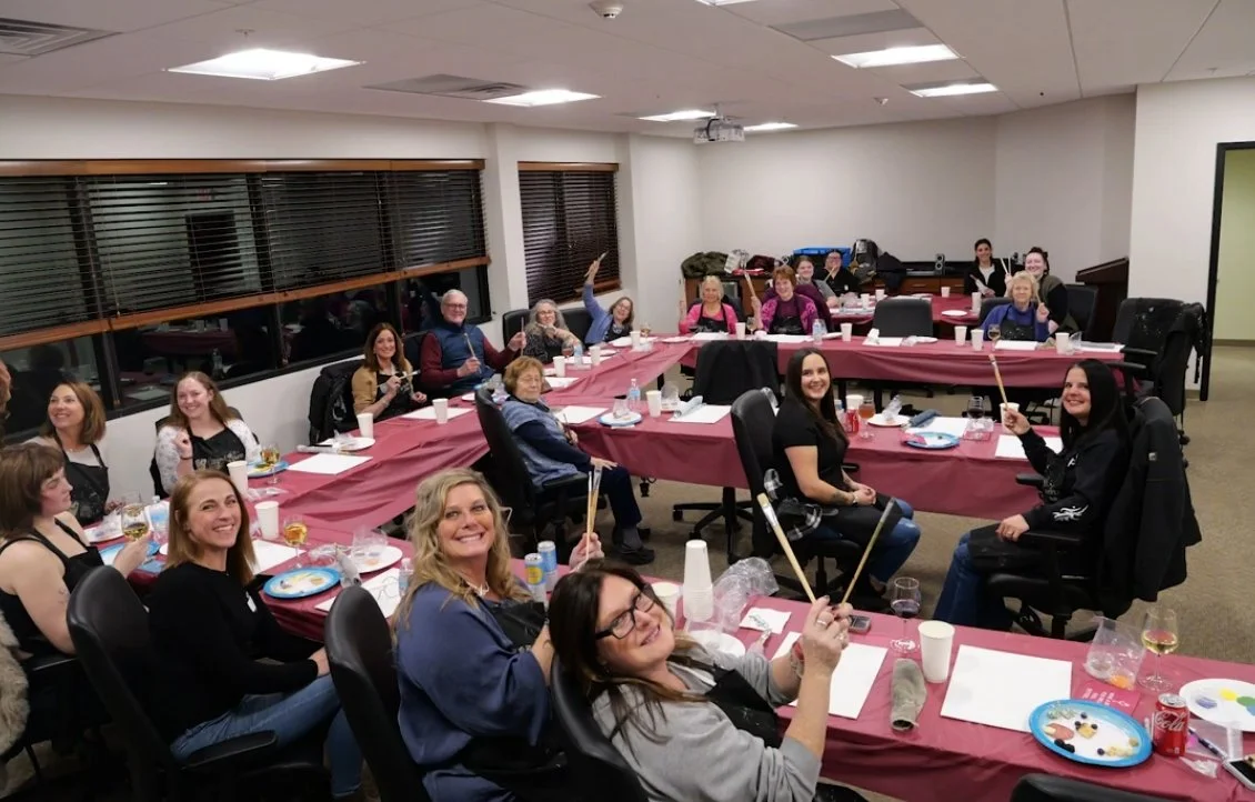 Group of women gathered in a conference room around U-shaped tables decorated with pink tablecloths, enjoying a celebration with snacks and drinks, some holding drumsticks.