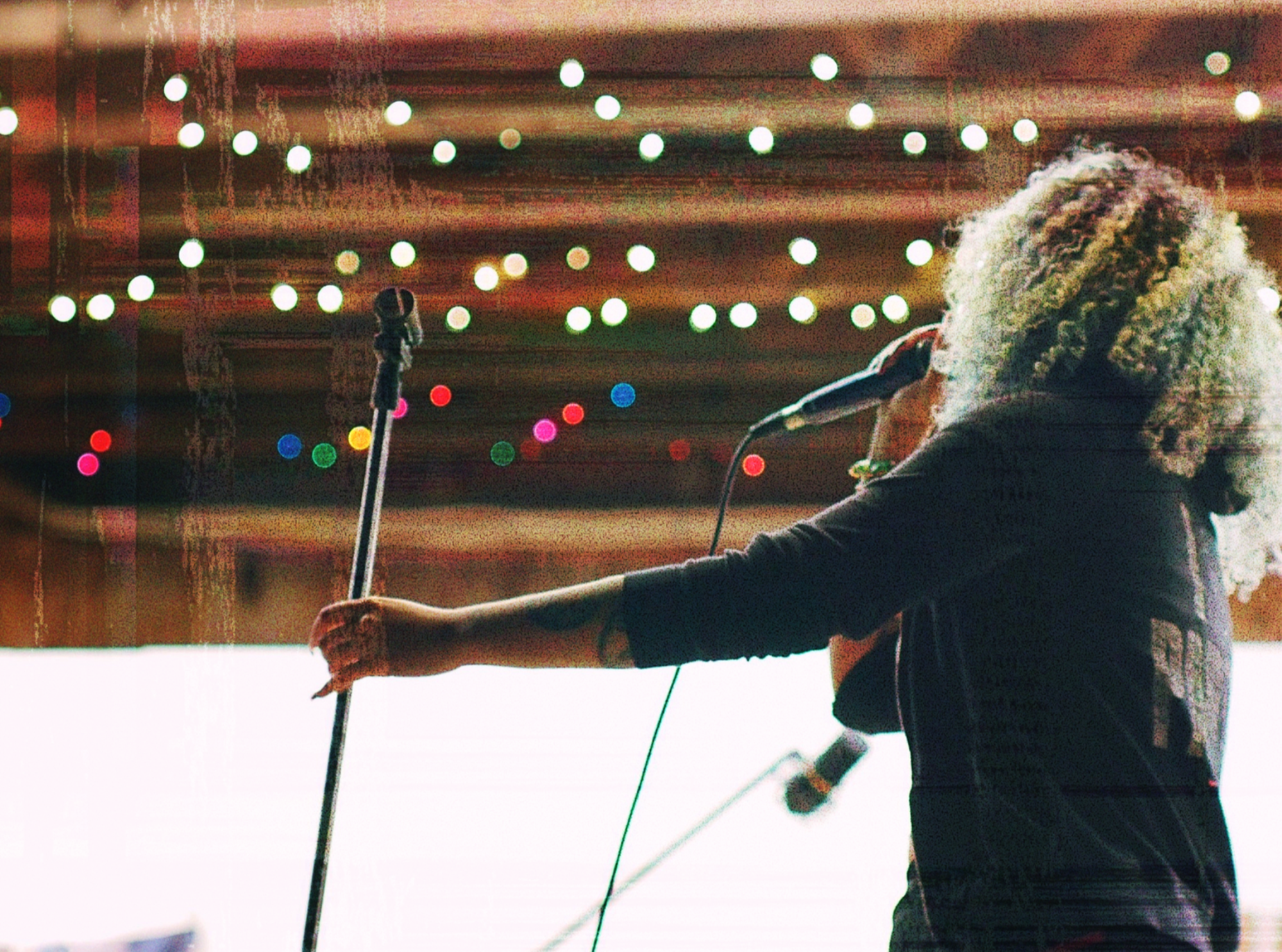 A woman with curly colorful hair singing into a microphone on stage, with colorful string lights hanging in the background.