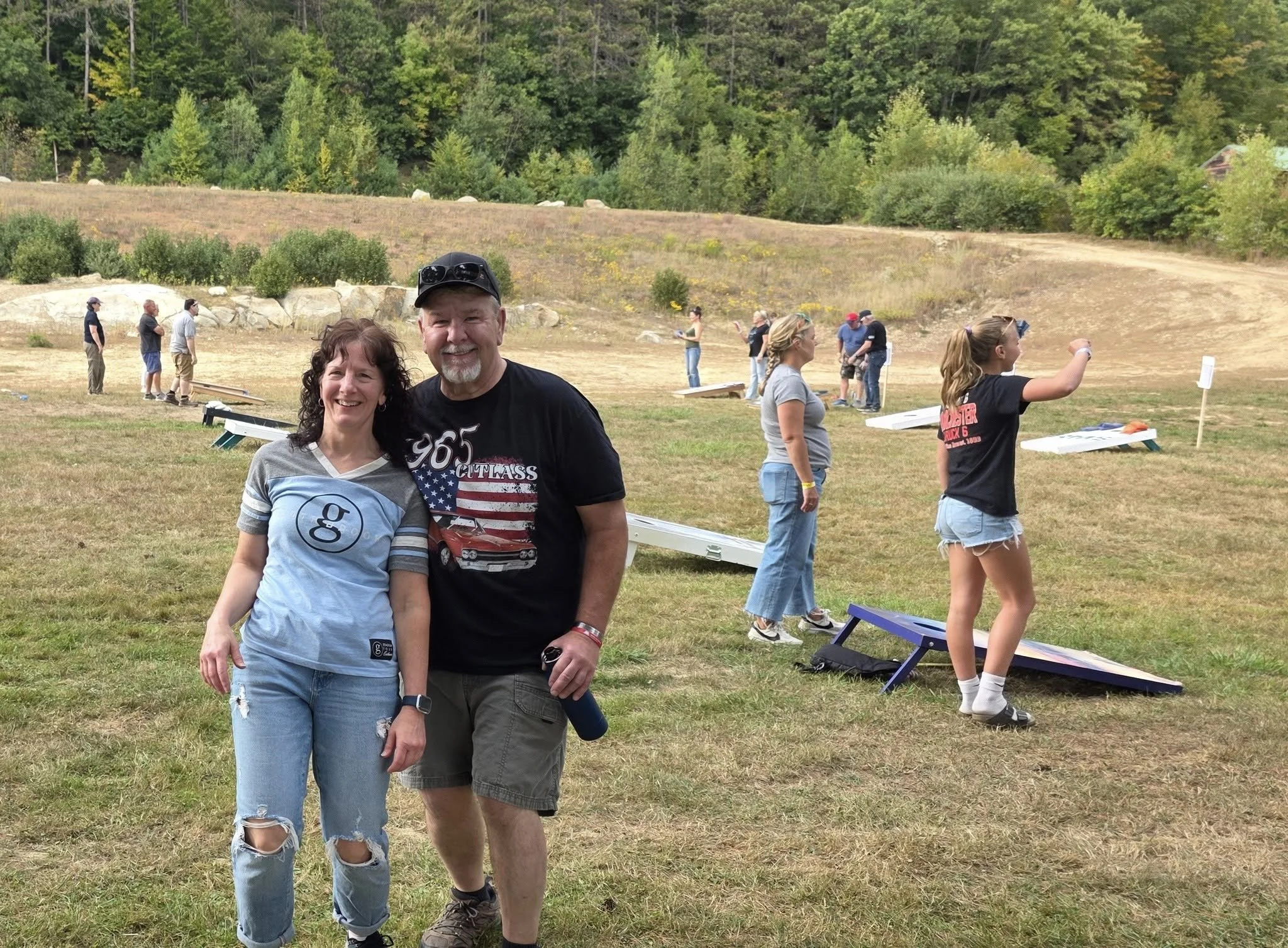 A man and a woman smiling and posing outdoors in front of a cornhole game at a grassy field with some trees and hills in the background. Several other people are seen in the background playing or watching the game.