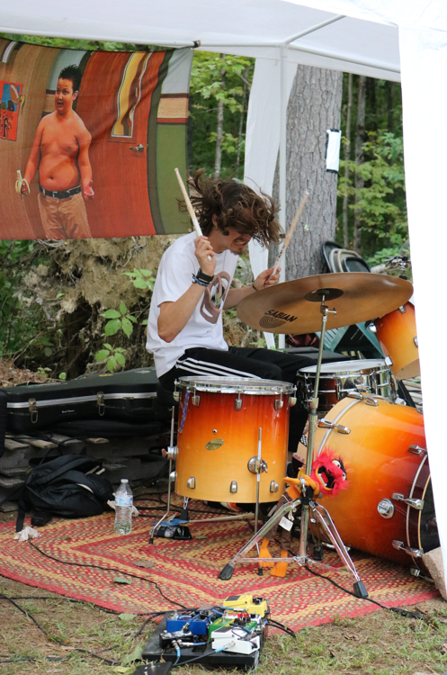 A young man with curly hair playing drums outdoors under a white canopy, with a colorful poster of a boy on the wall behind him, and trees in the background.