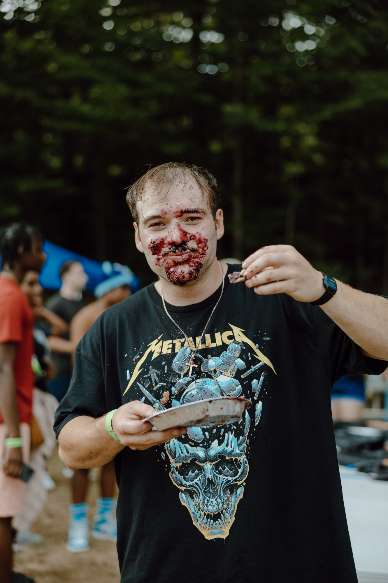 A man with a messy face, mostly covered in red cherry pie, holding a plate and a piece of pie, wearing a black t-shirt, at an outdoor gathering.